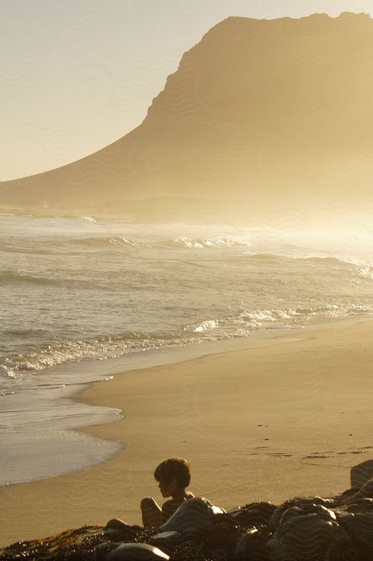 Landscape of a beach with the sea coast next to it and a boy sitting next to rocks.