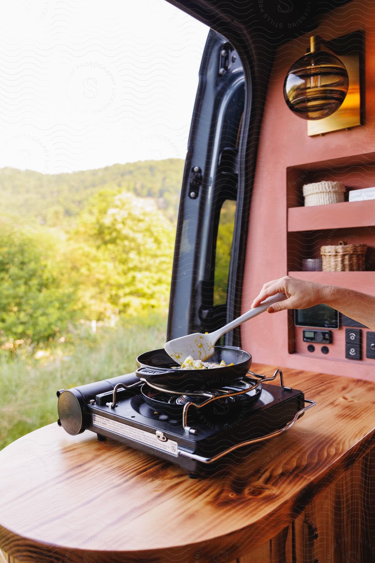 Someone is cooking on a portable stove in a vehicle, with the door open to reveal a panoramic view of the outdoors.