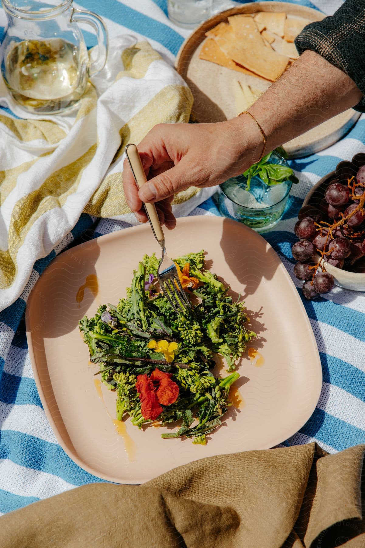 A man's hand holding a fork on a plate with vegetables on top of a towel and around a jug and a bowl with purple grapes.