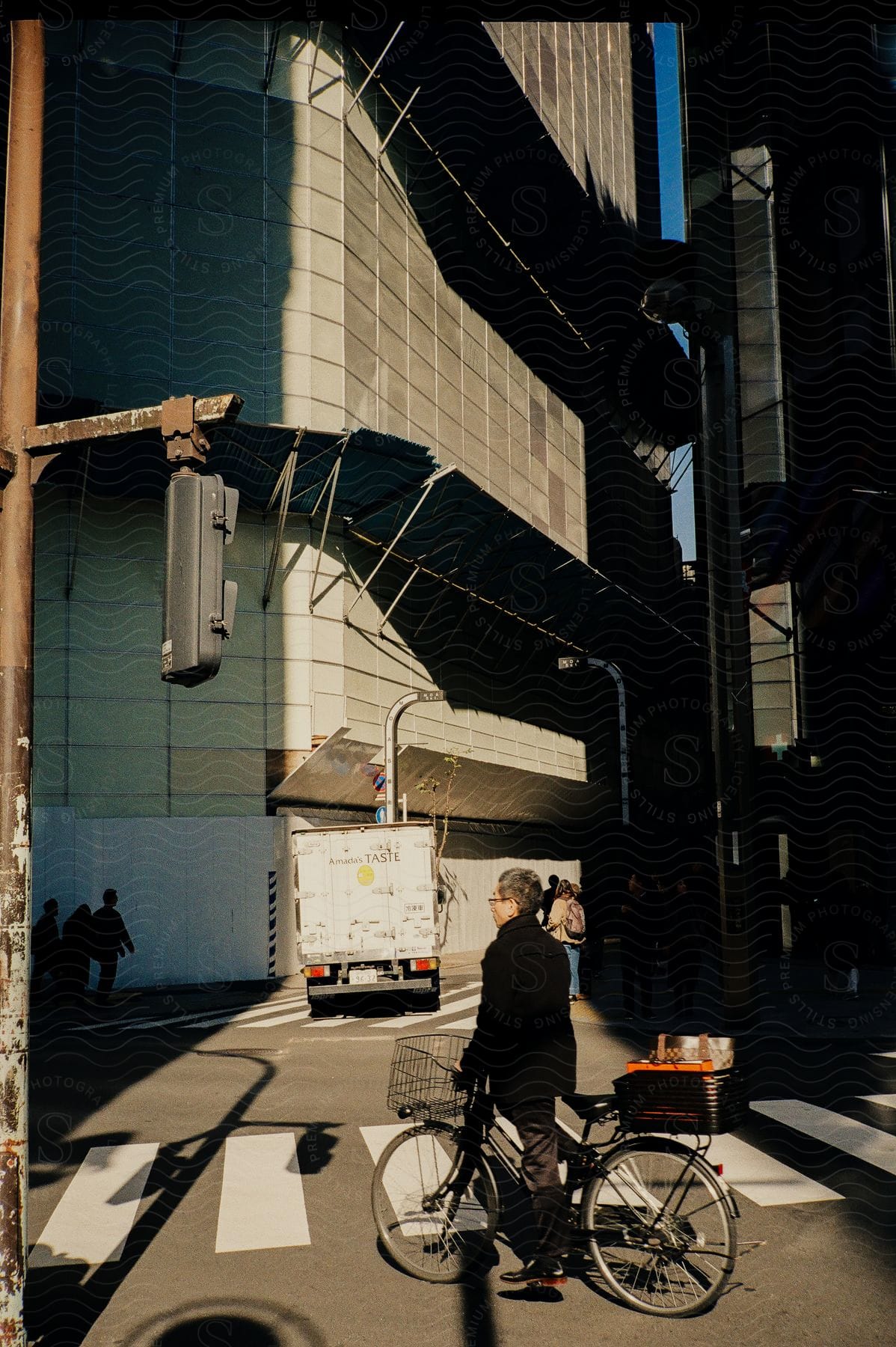 Man on a bicycle crossing a street as people stand near a building and a delivery truck