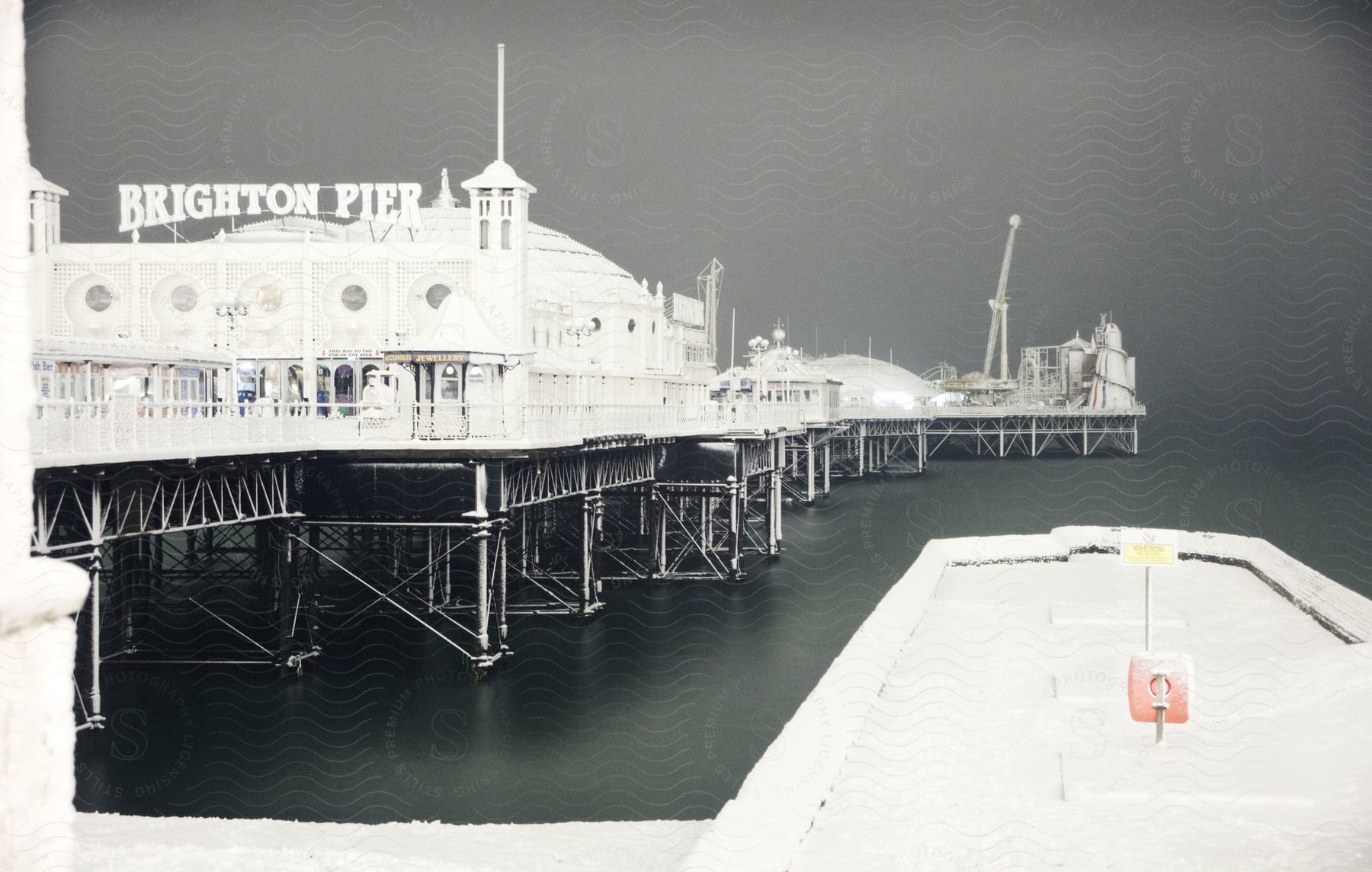 A night scene of the snow-covered Brighton Pier, illuminated against the dark sky. The pier, with its classic architecture, is supported by dark pillars.