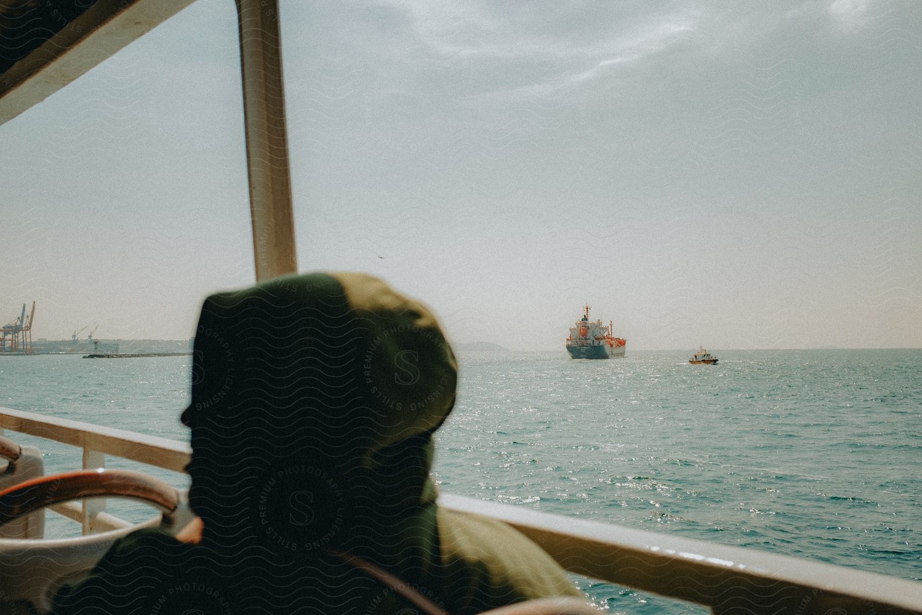 Person wearing a hooded jacket sitting on a boat as ships sail in the distance