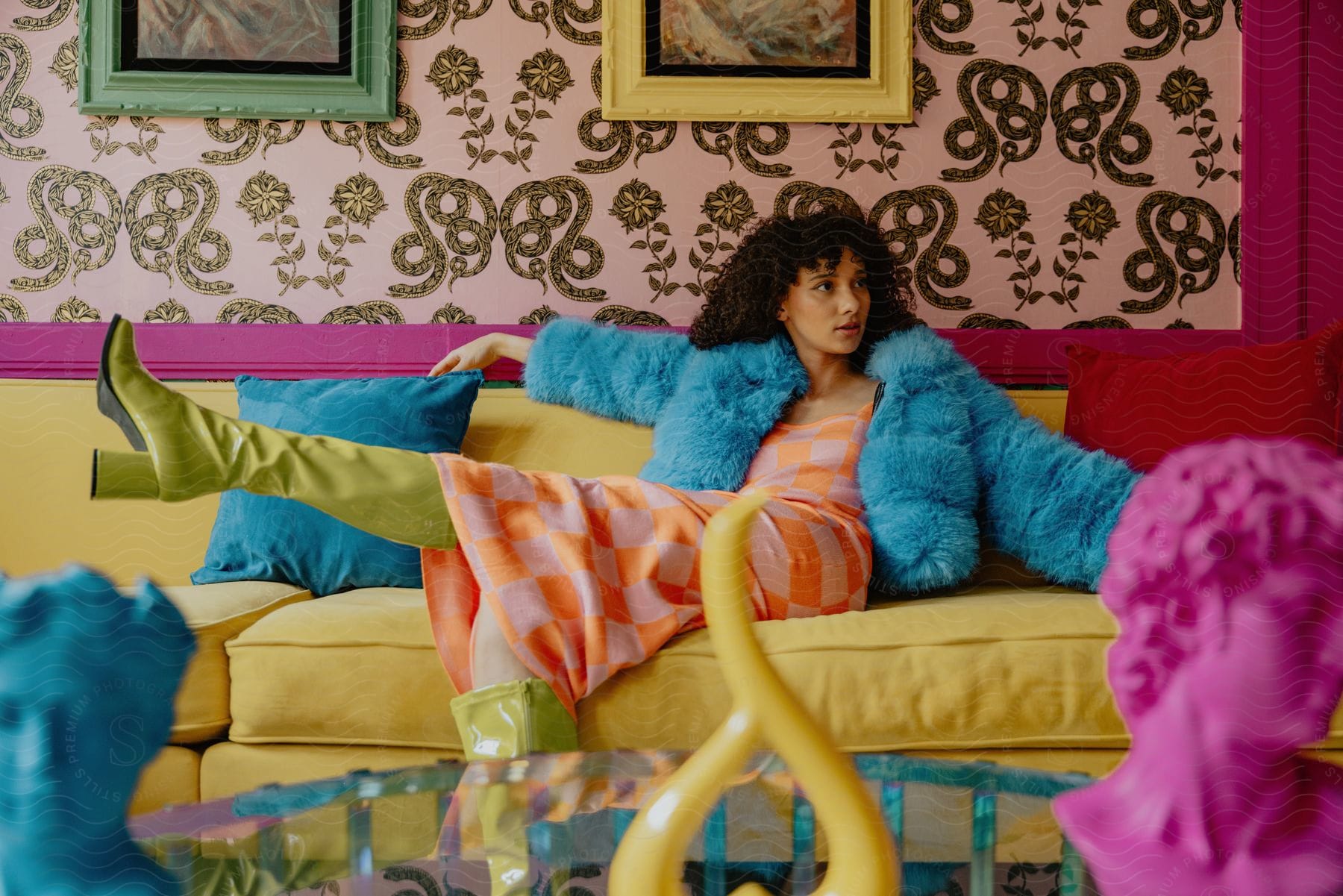 Woman reclining on a yellow sofa with blue cushion in a vibrantly colored room, wearing a blue furry coat and green boots.