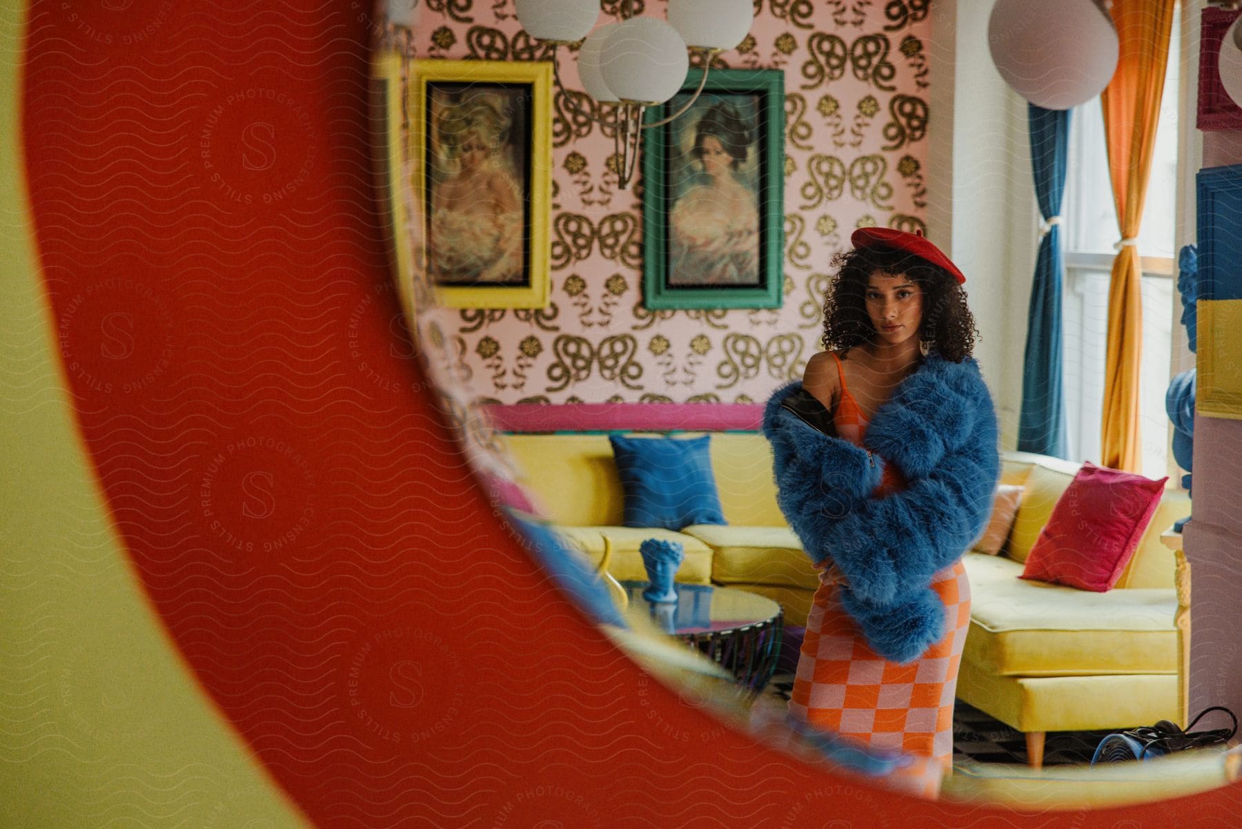Reflection in a mirror of a young woman wearing a beret and a blue furry coat posing in a garishly colored room