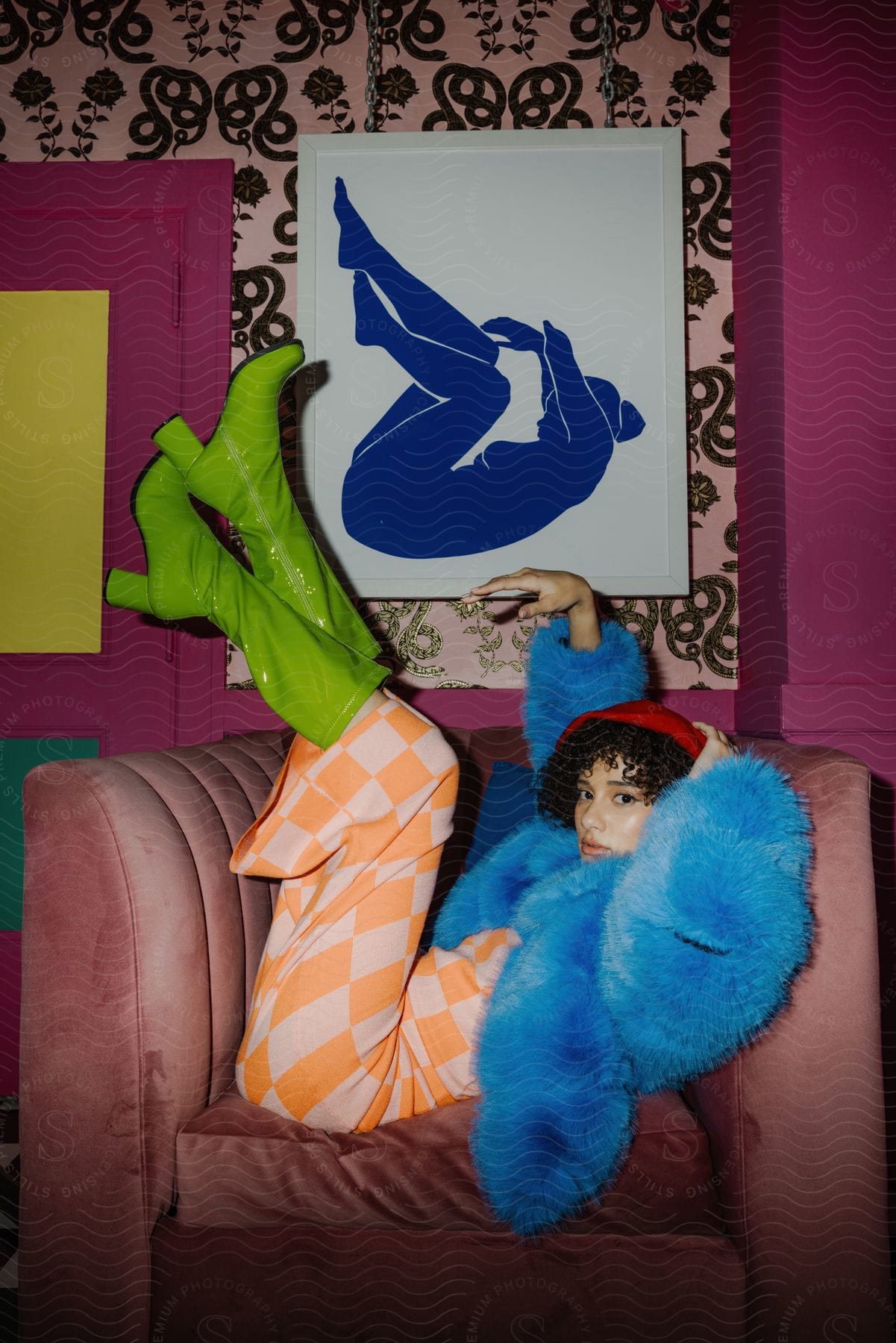 Woman reclining on a pink armchair with blue furry coat and green boots, orange and white striped dress with blue silhouette art behind her.