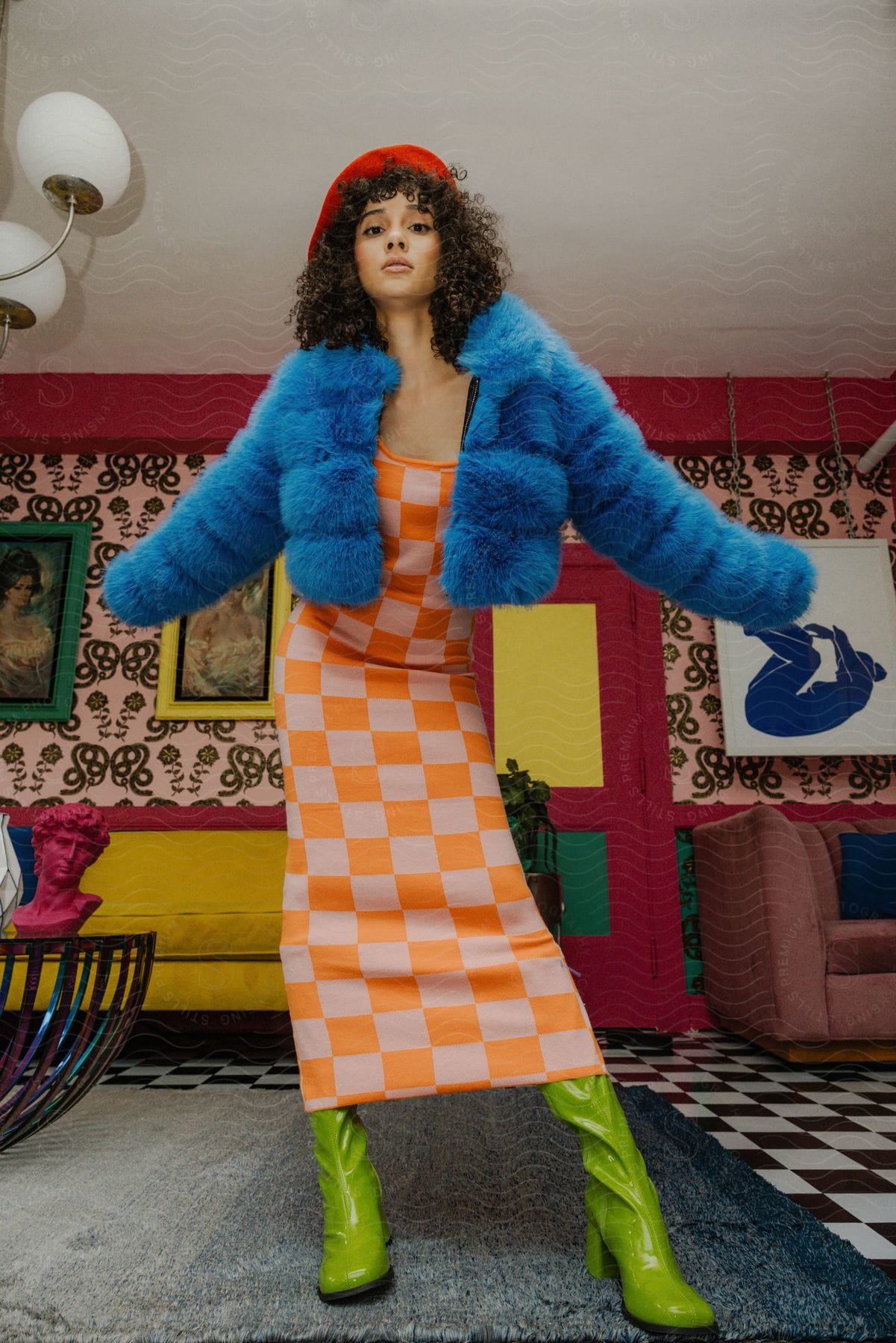 A young woman stands with her arms out while modeling a blue coat and checkerboard dress.