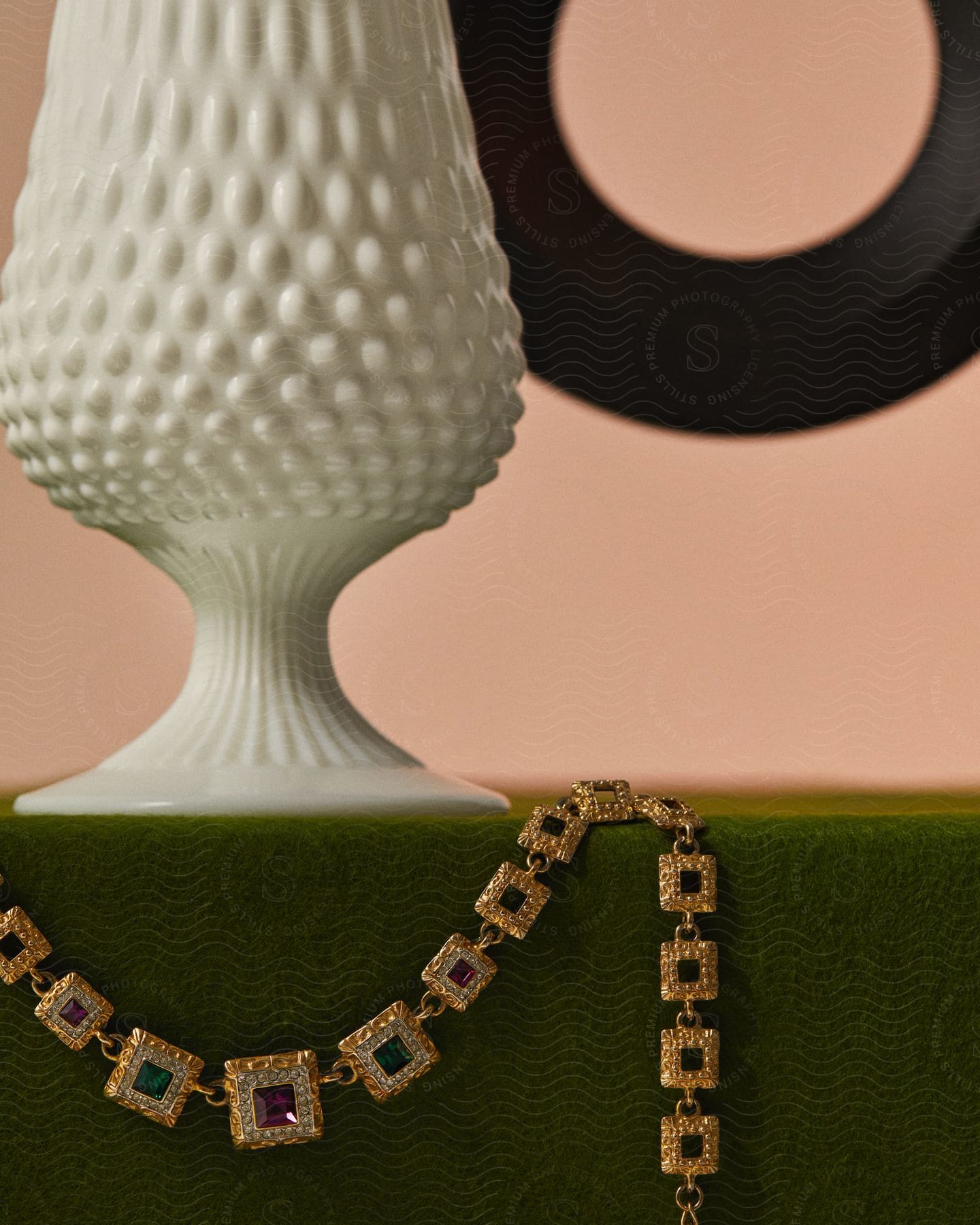 A gold one with colored gemstones is arranged on a green surface, next to a white vase with an intricate texture and a peach-colored wall with part of a circular work of art visible in the top right corner.