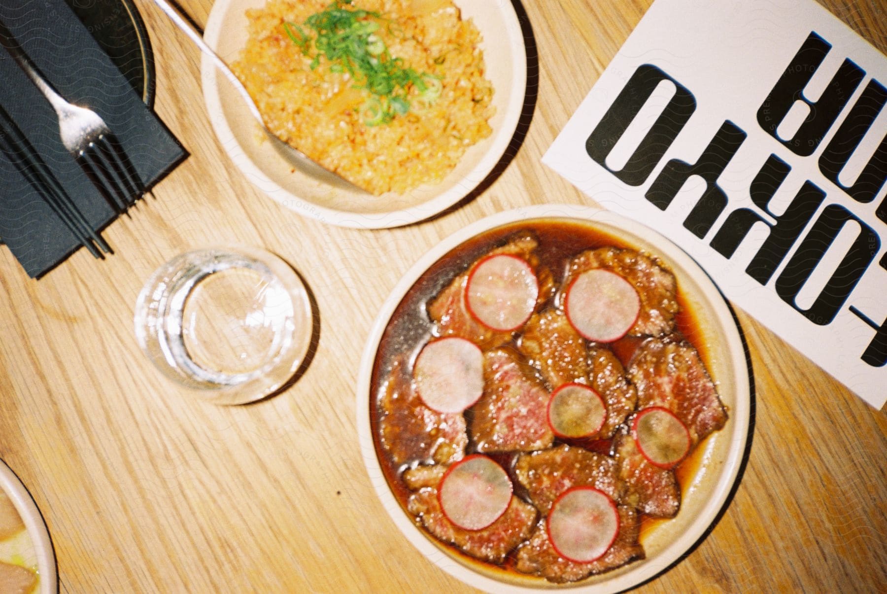 Top-down view of a dining table with a bowl of fried rice garnished with green onions, a bowl of beef with radish slices, an empty glass, black chopsticks, and a magazine with bold black and white lettering.