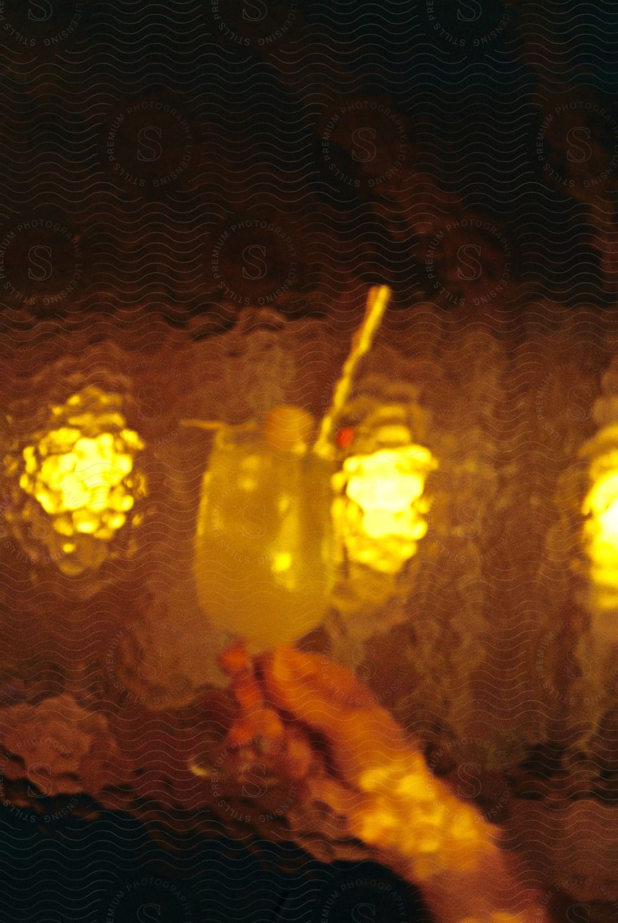 Hand holding a glass of a yellowish beverage with a straw and fruit garnish, reflected on a textured amber surface.
