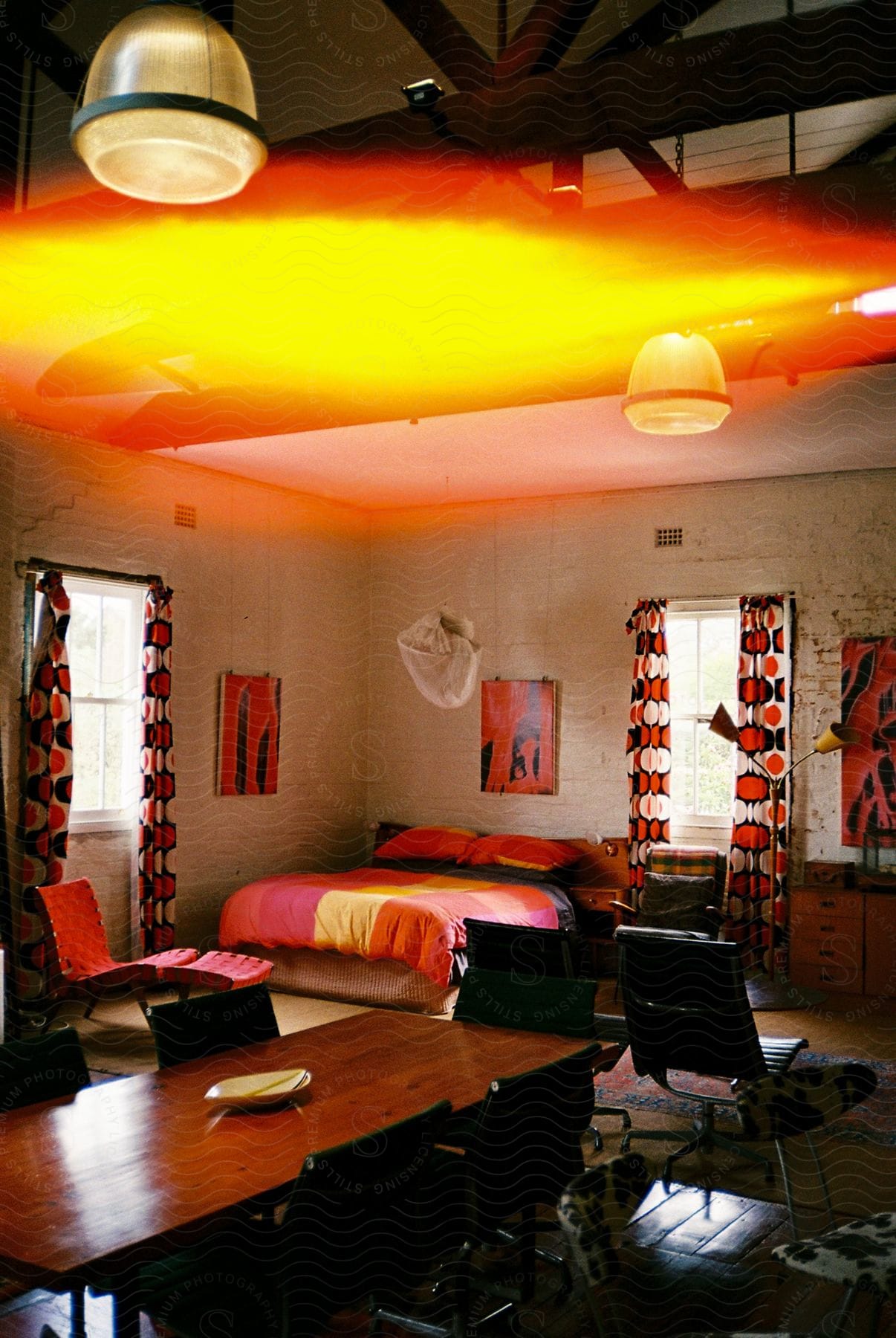 Interior of a bedroom with bright orange lighting, featuring a bed with red and orange bedding and curtains with a circular pattern. A wooden desk with chairs and a ceiling lamp is in the foreground.