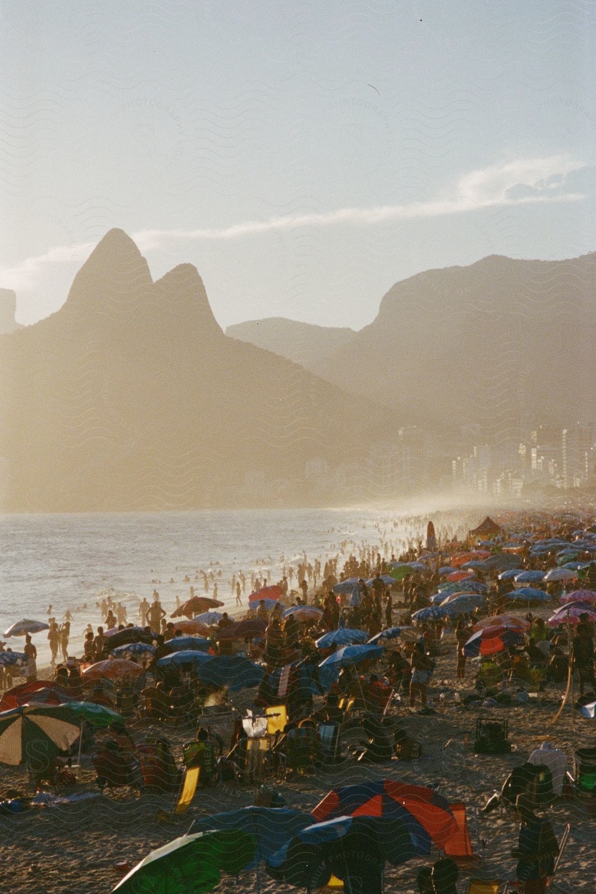 Crowded beach scene with numerous umbrellas at sunset with mountain silhouettes in the background.