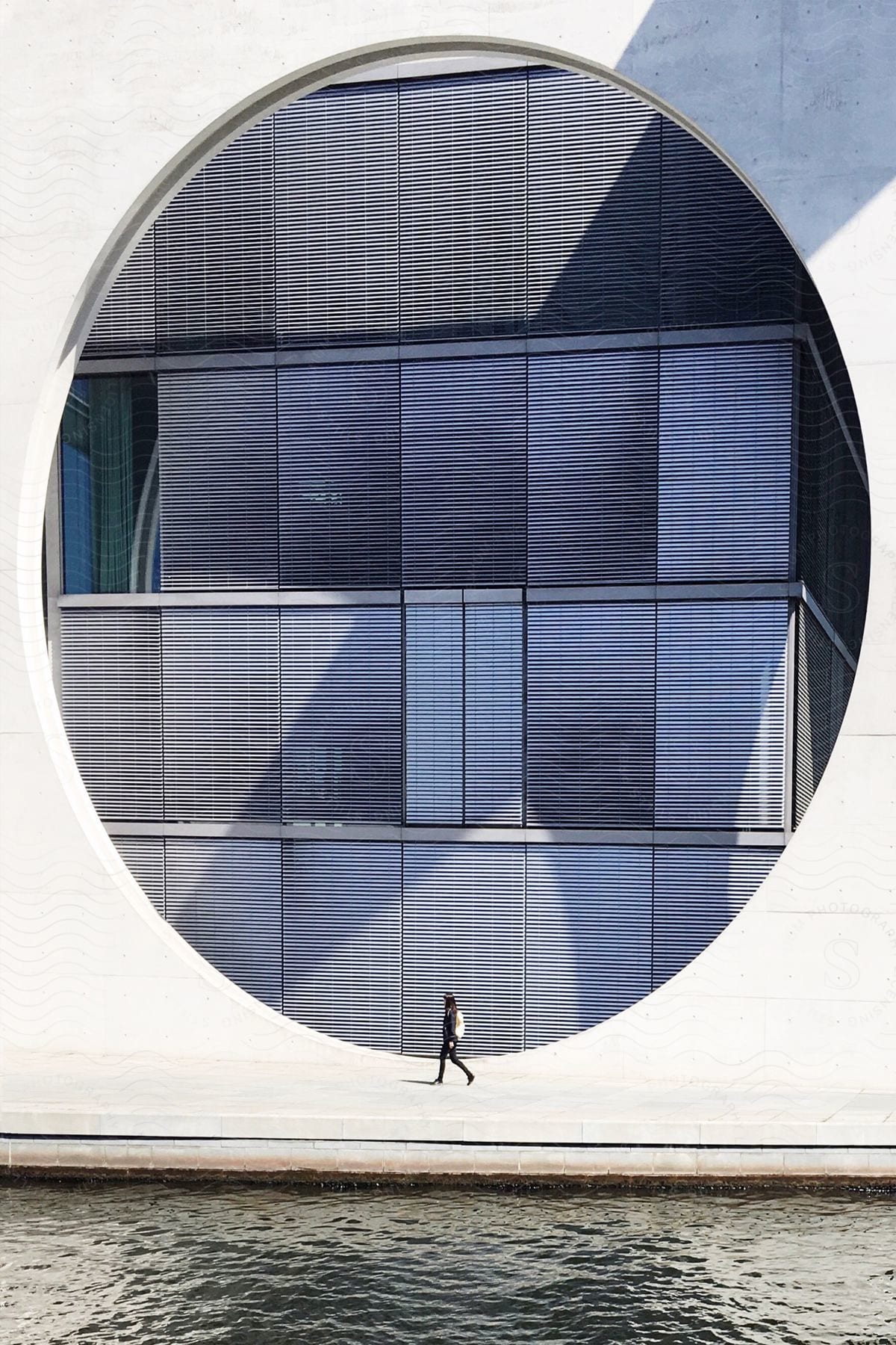 Person walking past a modern building with a circular facade overlooking water.