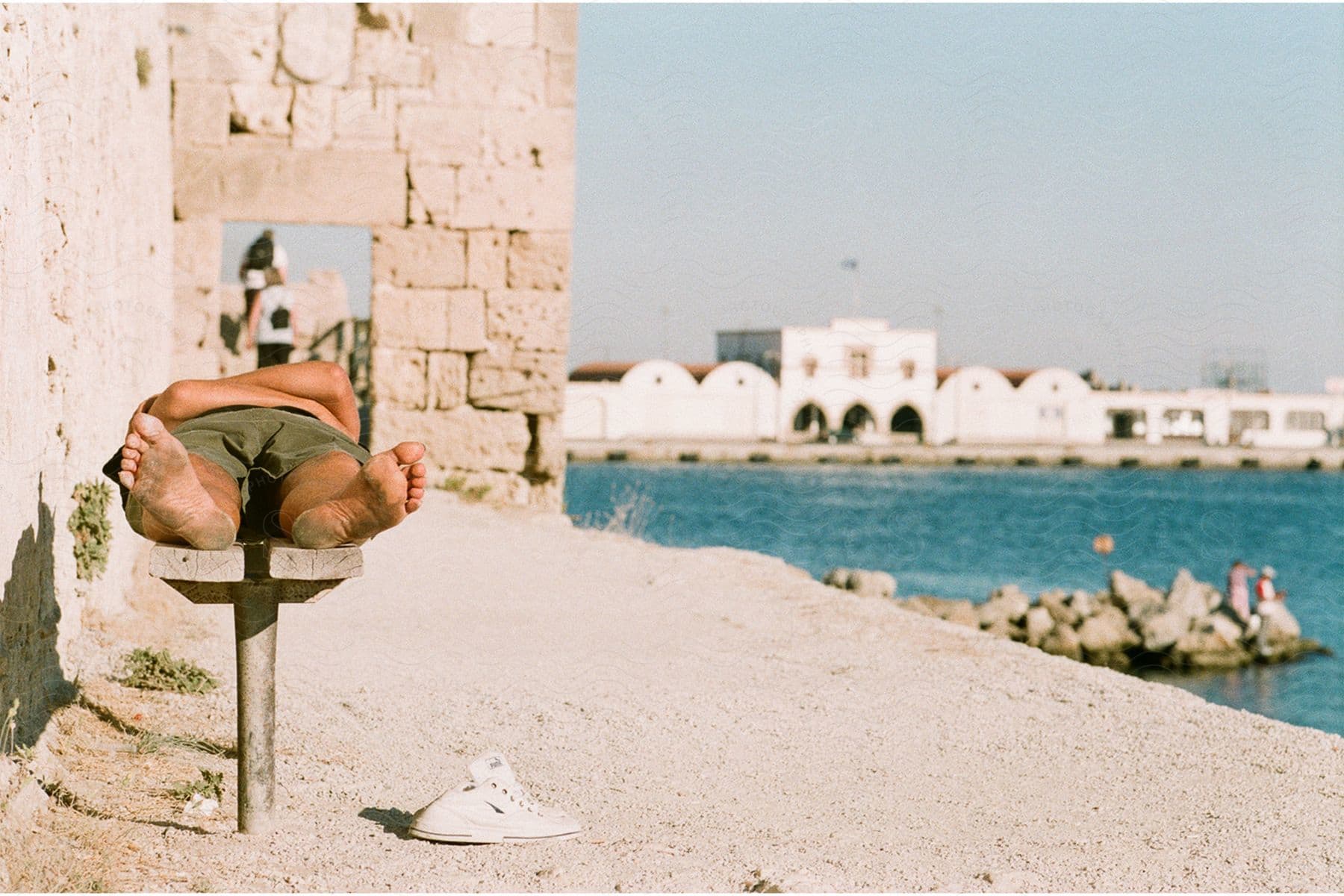 Barefoot man lying on a bench near a building along the coast