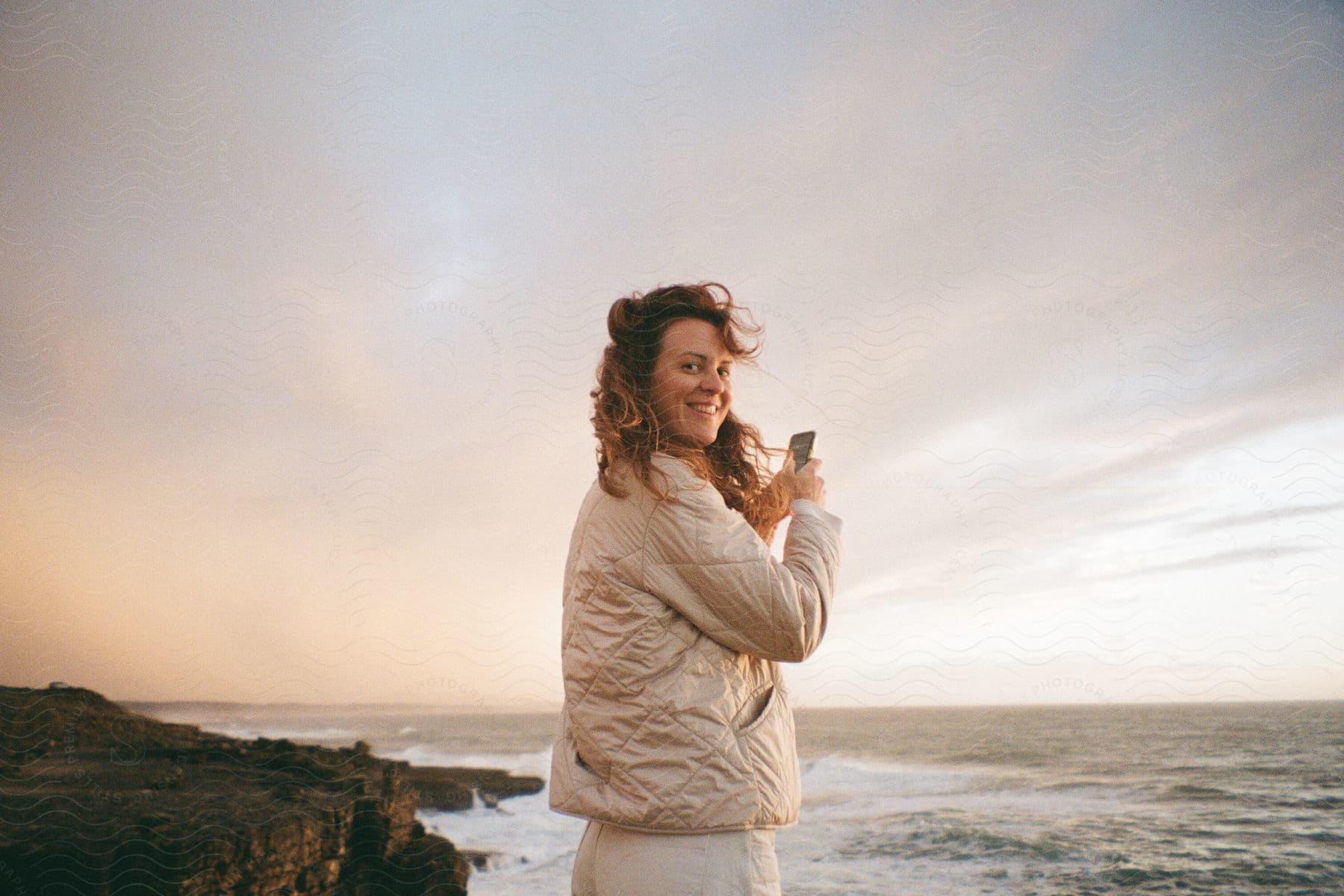 Woman standing along the coast holding her phone as she turns and smiles