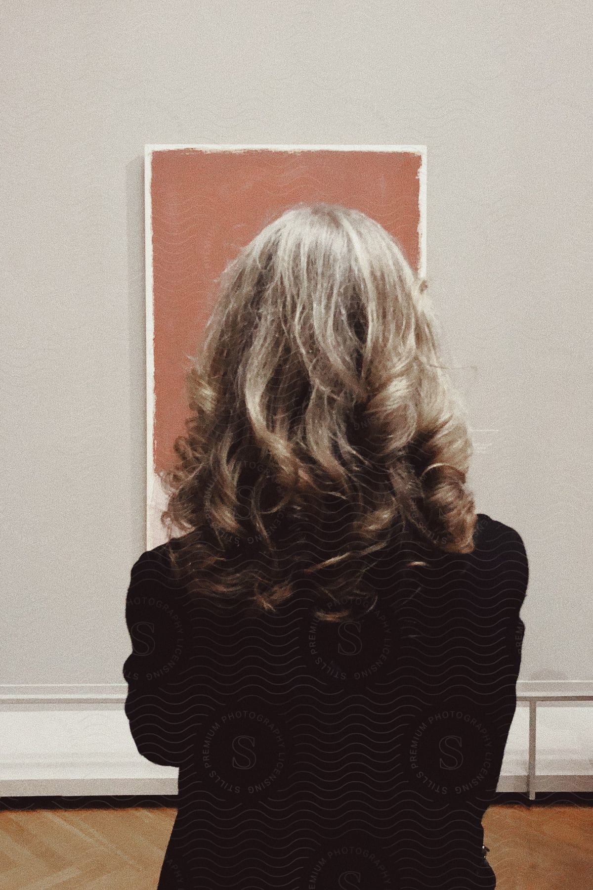 A blonde woman with back to the camera looks at a modern art painting in a gallery.