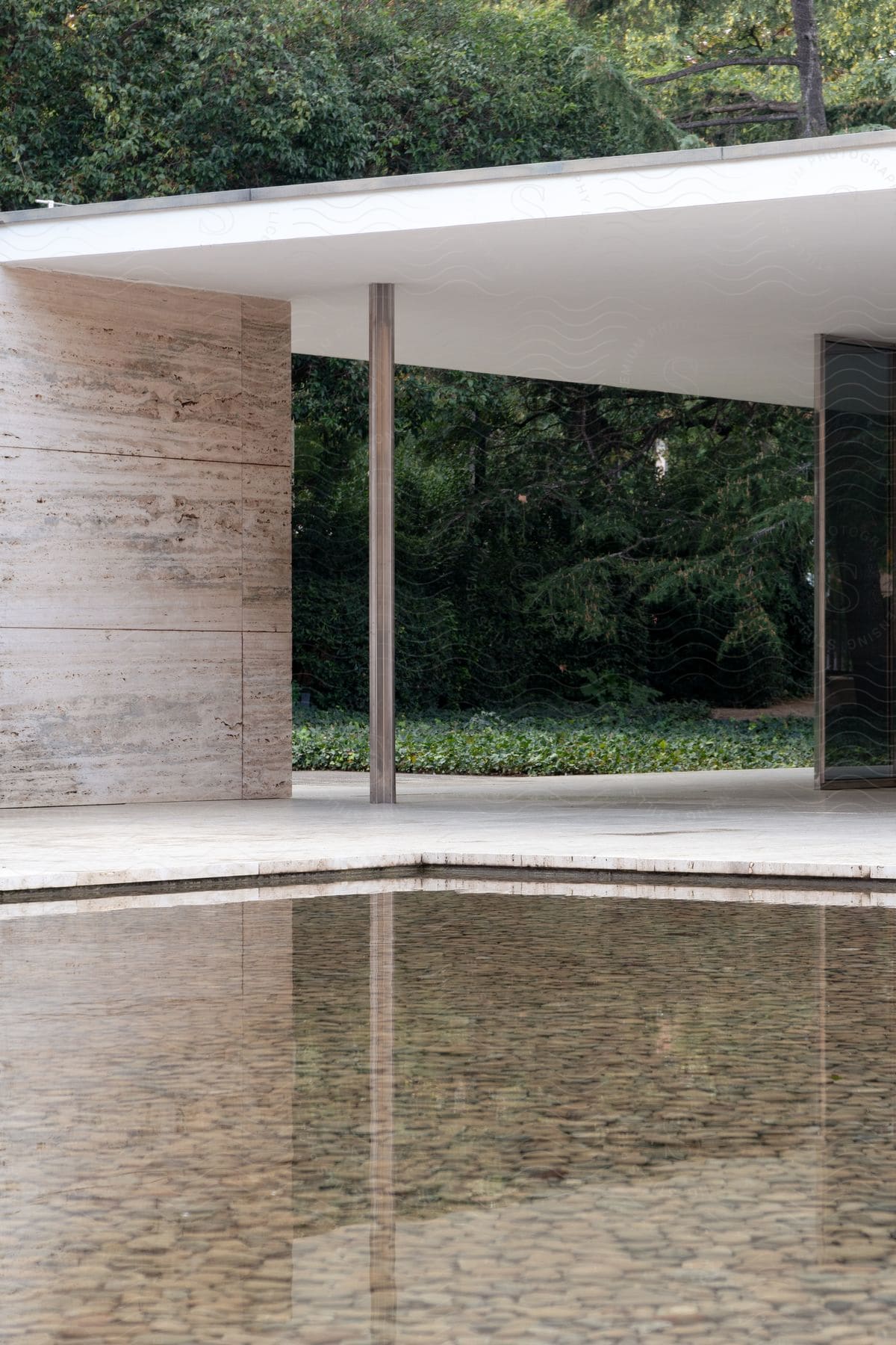 Modern architecture with a flat roof, supported by a column, overlooking a reflecting pool with a backdrop of trees.