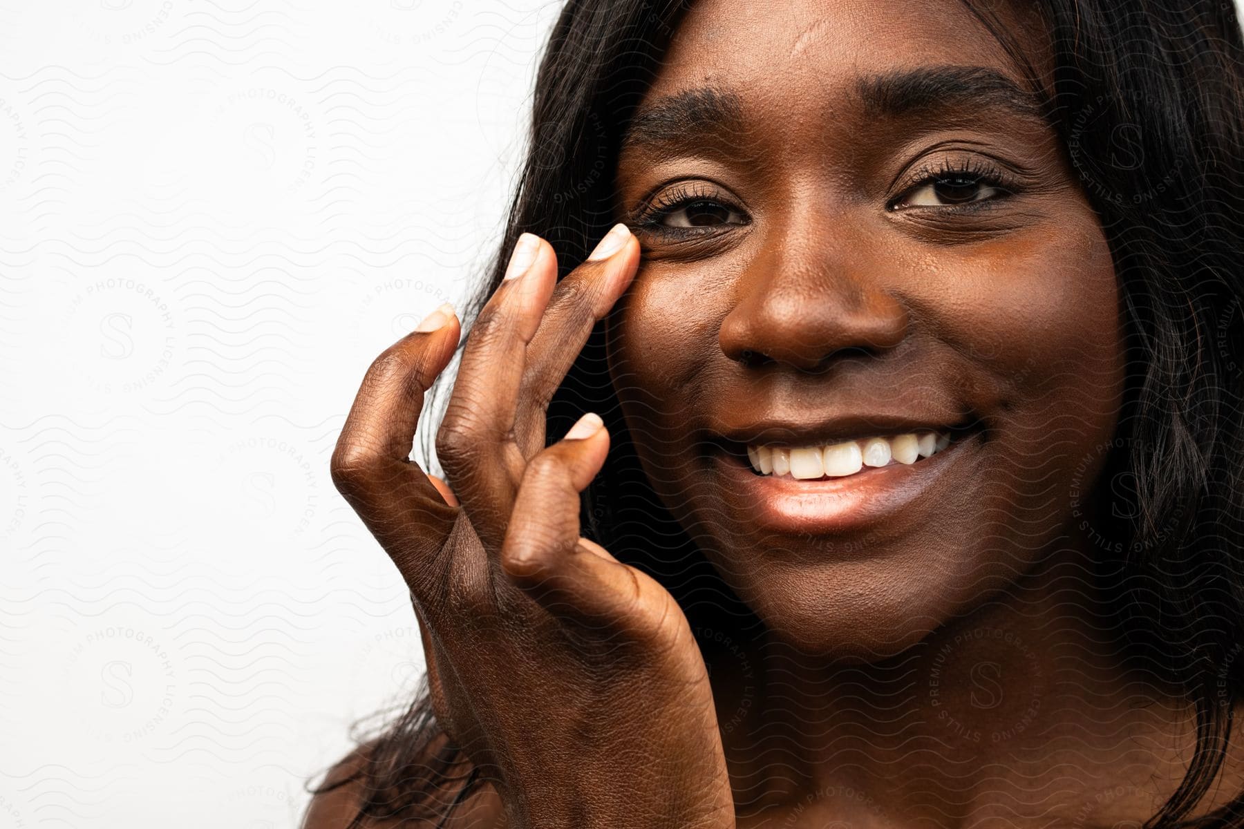 Close-up of a smiling woman applying skincare to her cheek, light background.