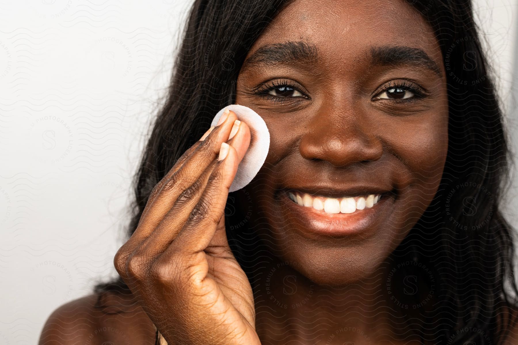 Face of a young smiling woman using a makeup removal cotton pad.