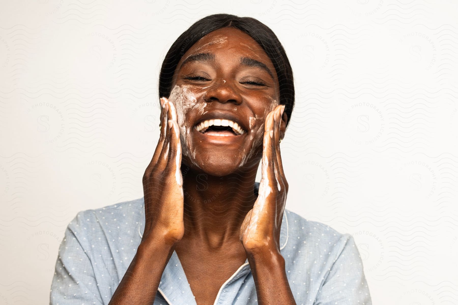 A black woman laughs as she rubs facial cream onto her face, her hands moving in gentle motions across her skin.