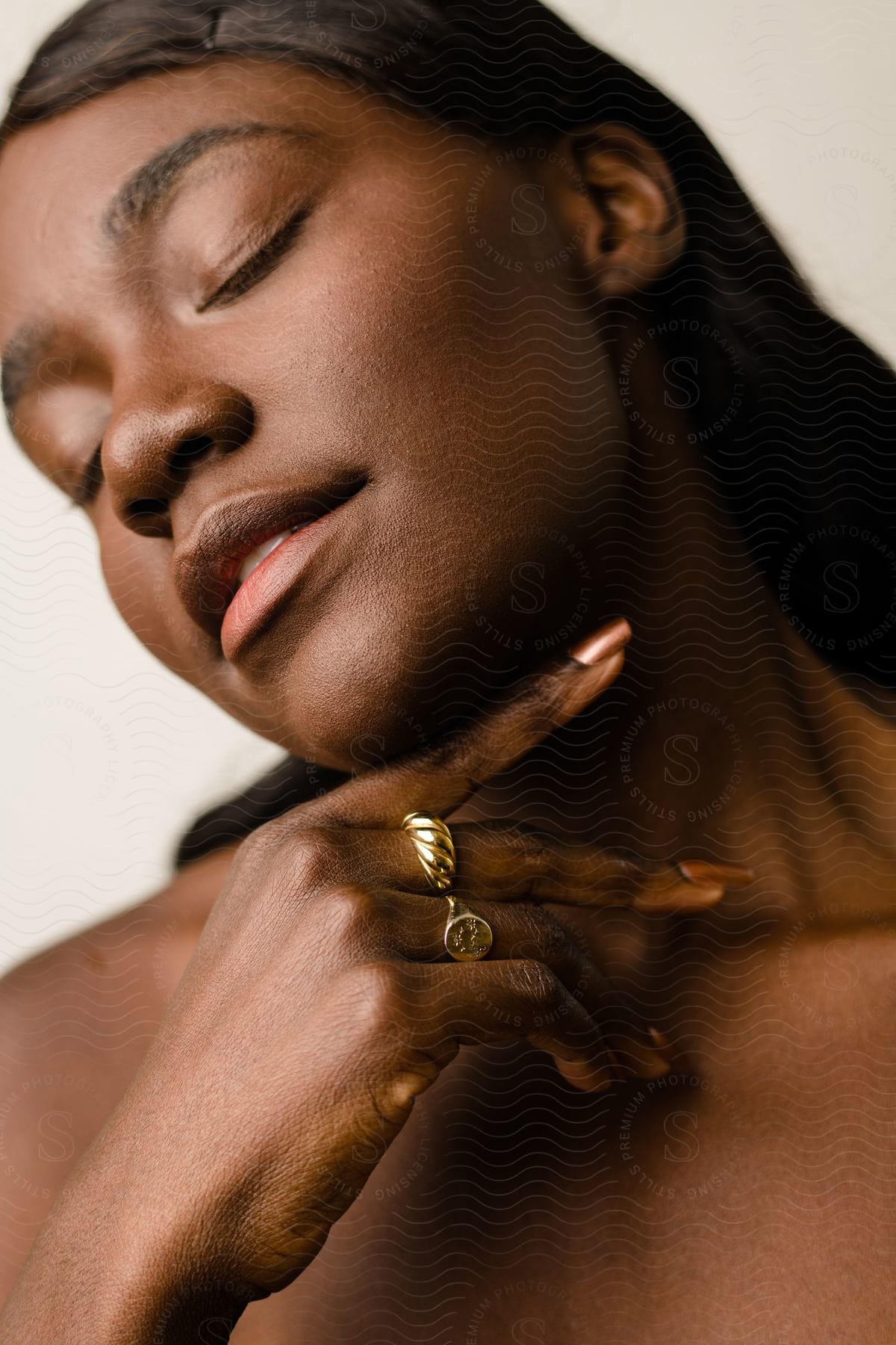 Young model with closed eyes and black hair posing with one hand on her chin and wearing golden rings.