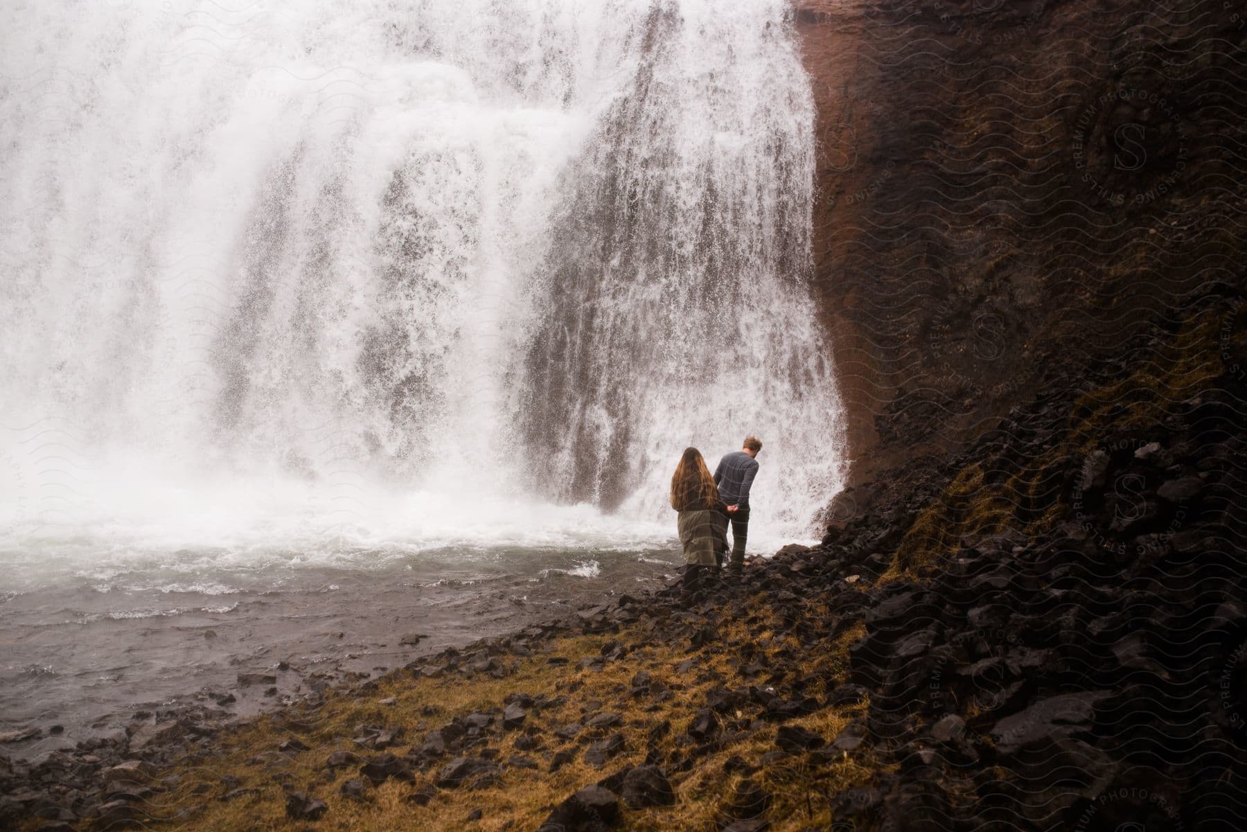 A couple watching a large waterfall. The waterfall is powerful, with white, foamy water falling from a considerable height.