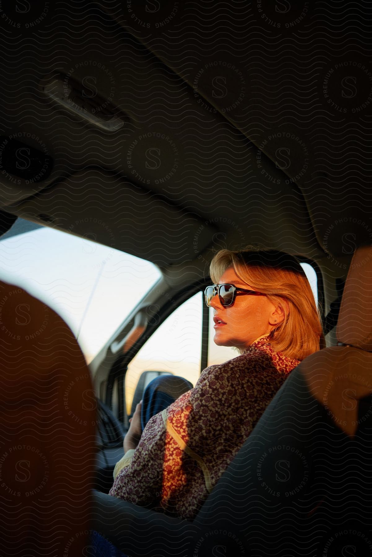 A blonde woman is wearing sunglasses while sitting in a car and she is having her face illuminated by natural light