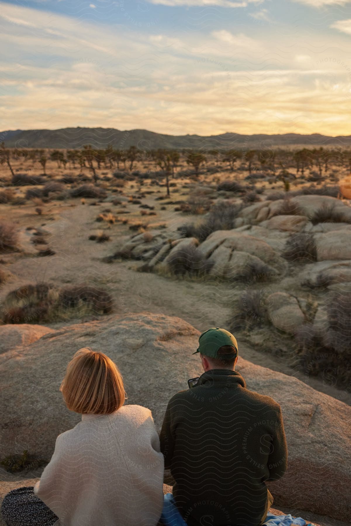 A young couple sits atop a rock, enjoying a romantic getaway in the valley as the sun sets. Rocks and trees surround them in the dry landscape.