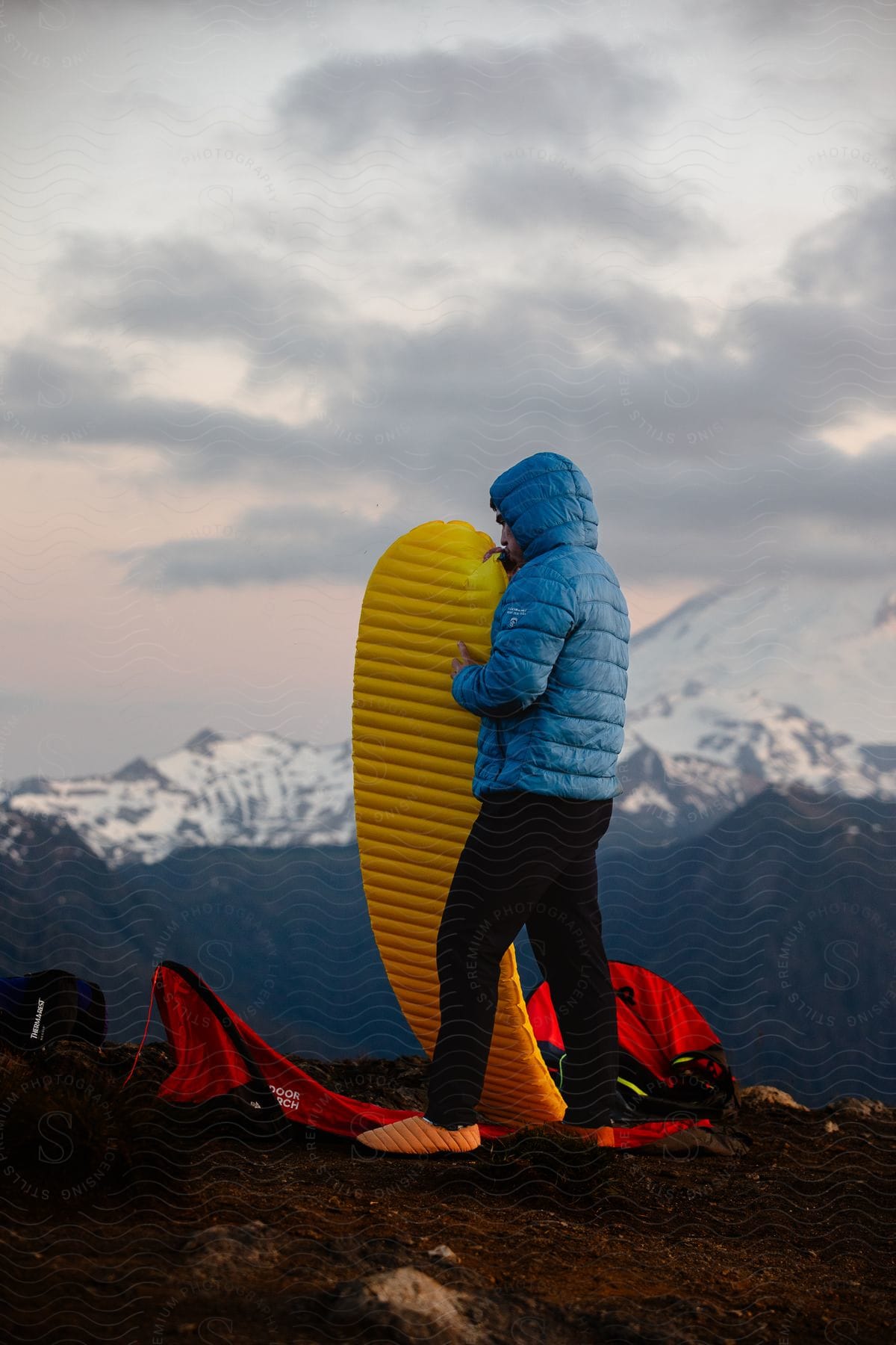 Individual in a blue jacket inflating a yellow sleeping pad on a mountain at dusk with snow-capped peaks in the distance and camping gear nearby.