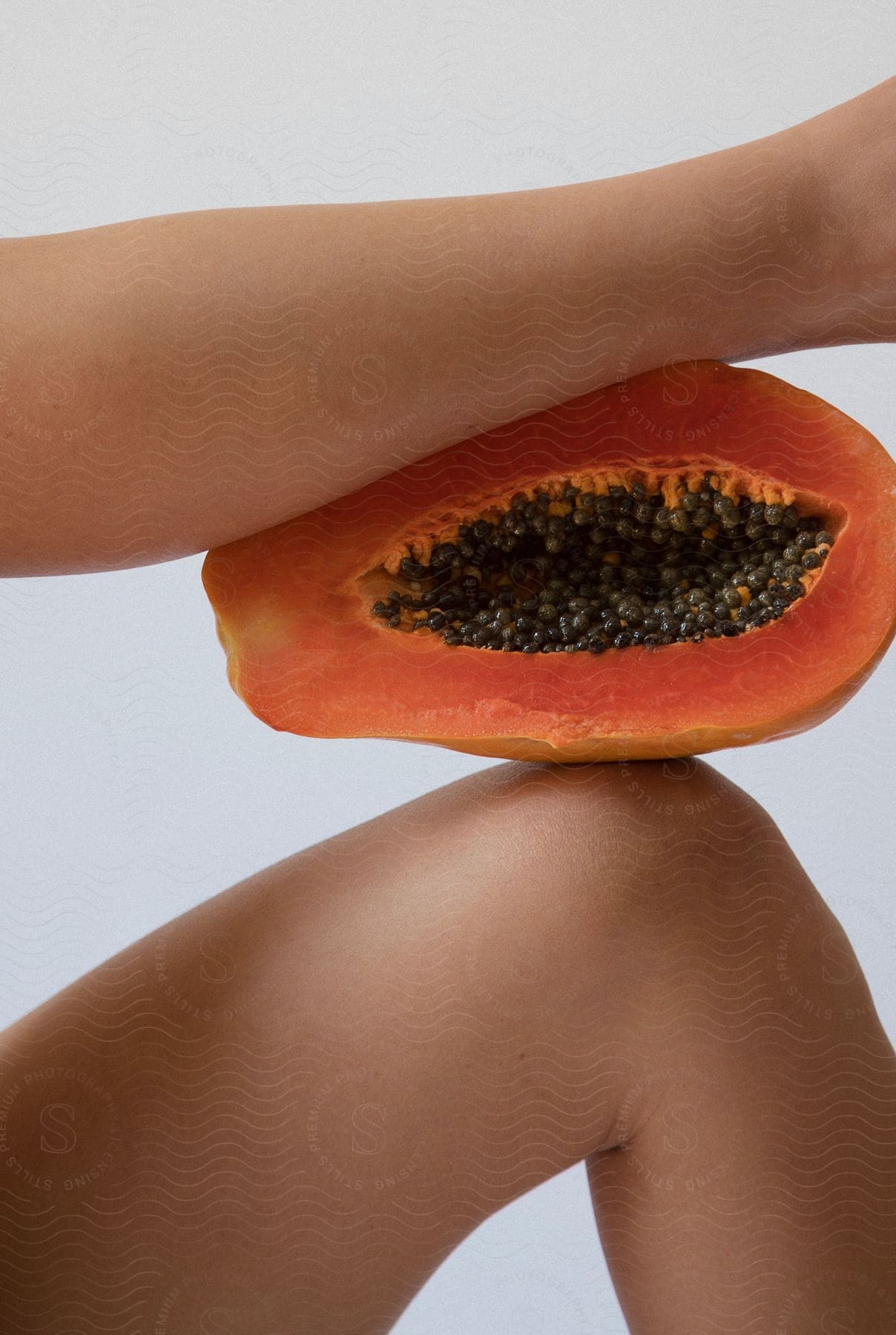 A halved papaya with seeds, resting between a person's bent arm and knee on a neutral background.