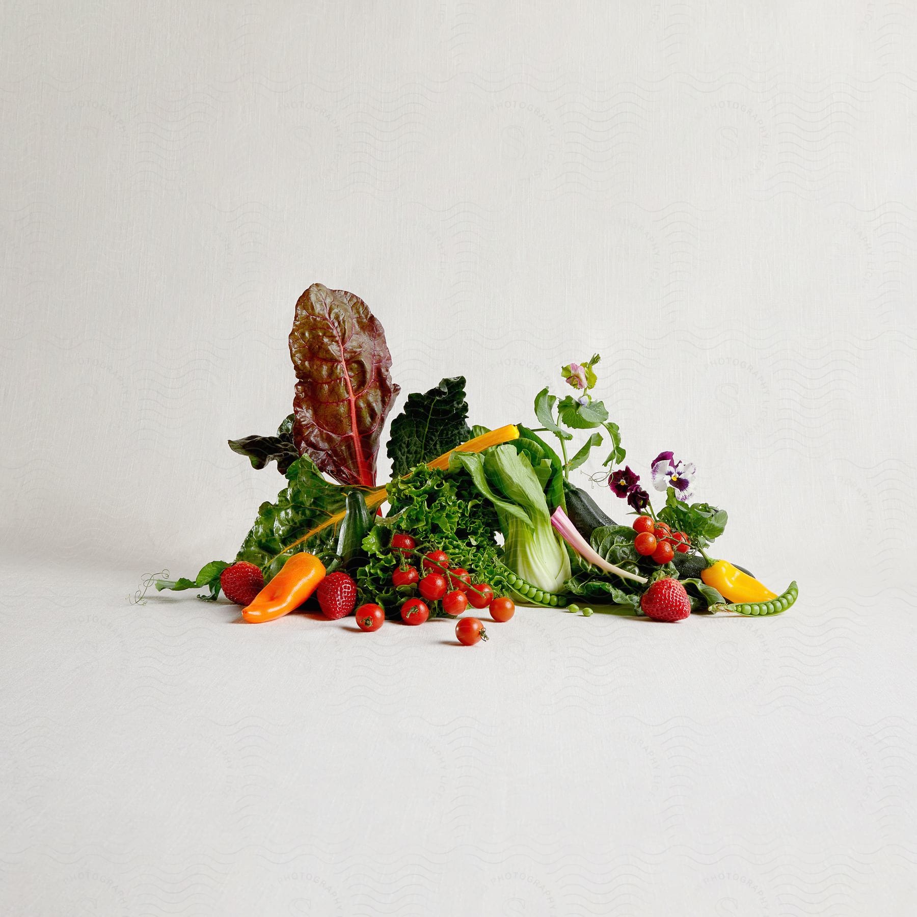 Diverse arrangement of colorful fresh vegetables and fruits, including leafy greens, tomatoes, peas, and strawberries, on a neutral backdrop.