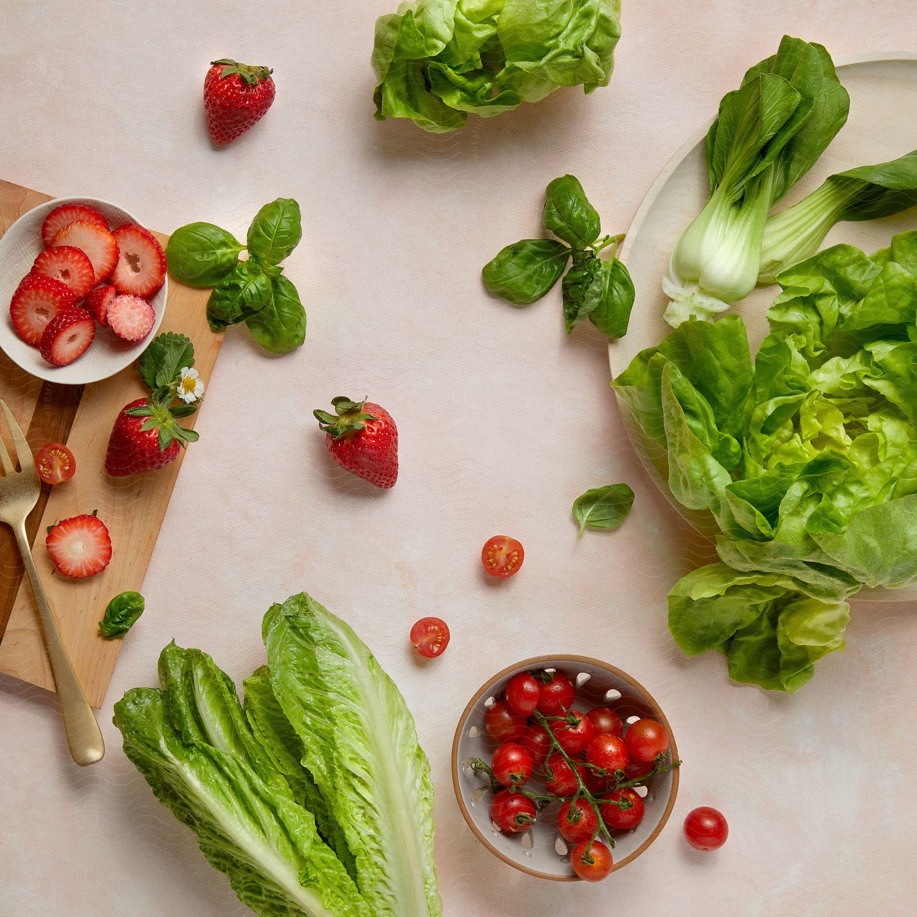 Various fresh vegetables and fruits arranged on a light pink surface; includes whole and sliced strawberries in a bowl, cherry tomatoes, lettuce, bok choy, and basil leaves.