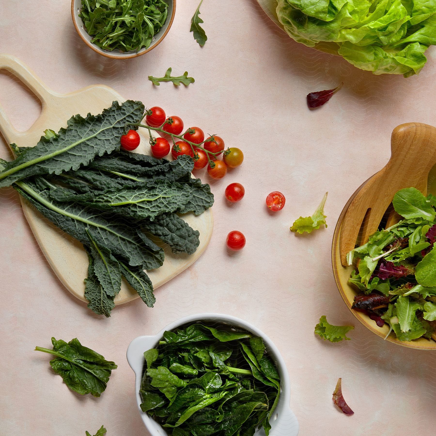 Kale on a cutting board, cherry tomatoes, mixed leaf salad in a wooden bowl, and spinach in a white bowl, all arranged on a pale pink surface.