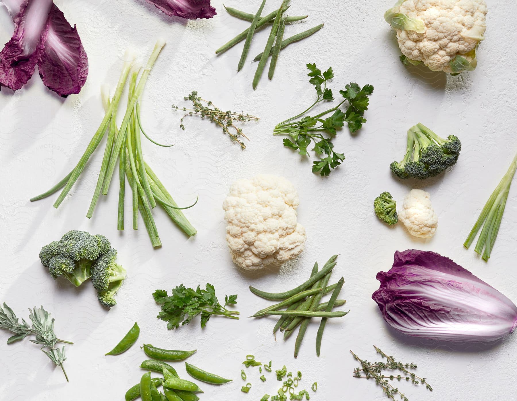 Various fresh vegetables, including broccoli, cauliflower, green beans, snap peas, and red lettuce, arranged on a white textured surface with scattered herbs.