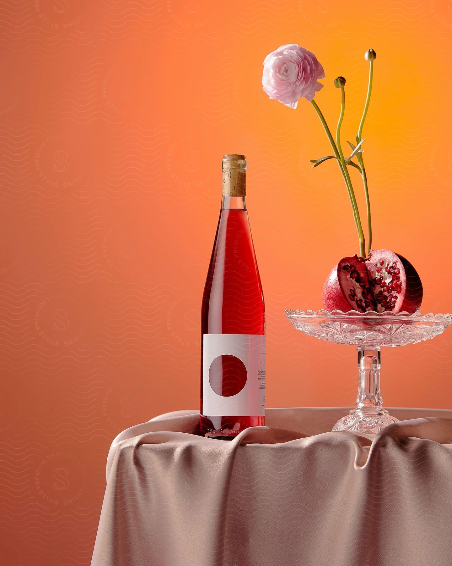 A red wine bottle sits next to a candy dish holding a cut open pomegranate with a carnation flower on a long stem, along with two other stems with flower buds, on a pink tablecloth with an orange-yellow background.