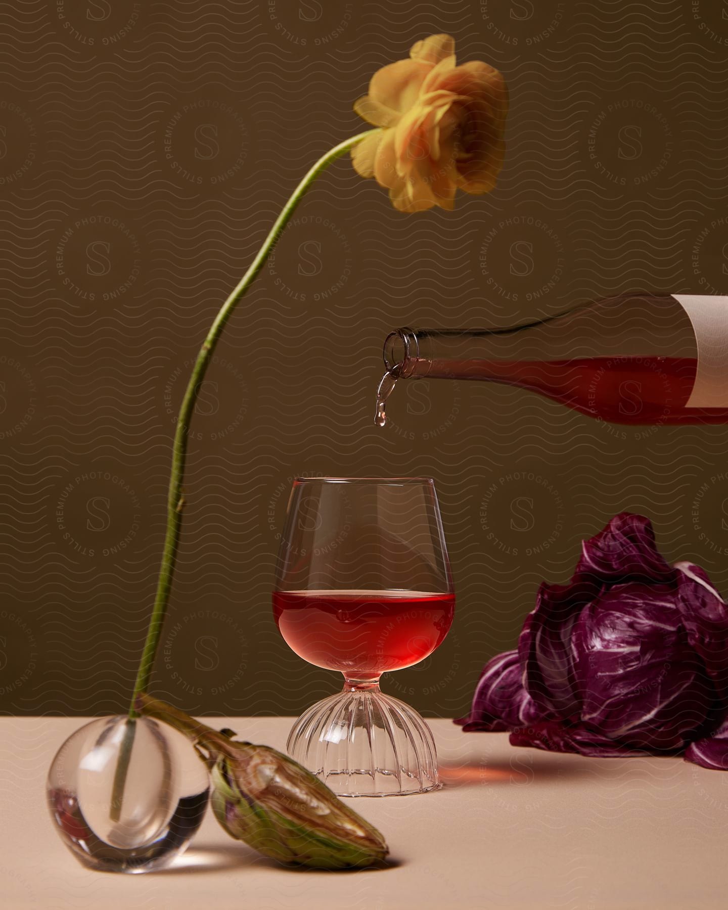 Red wine fills a glass, while a flower sits on a table with a bottle and another glass.