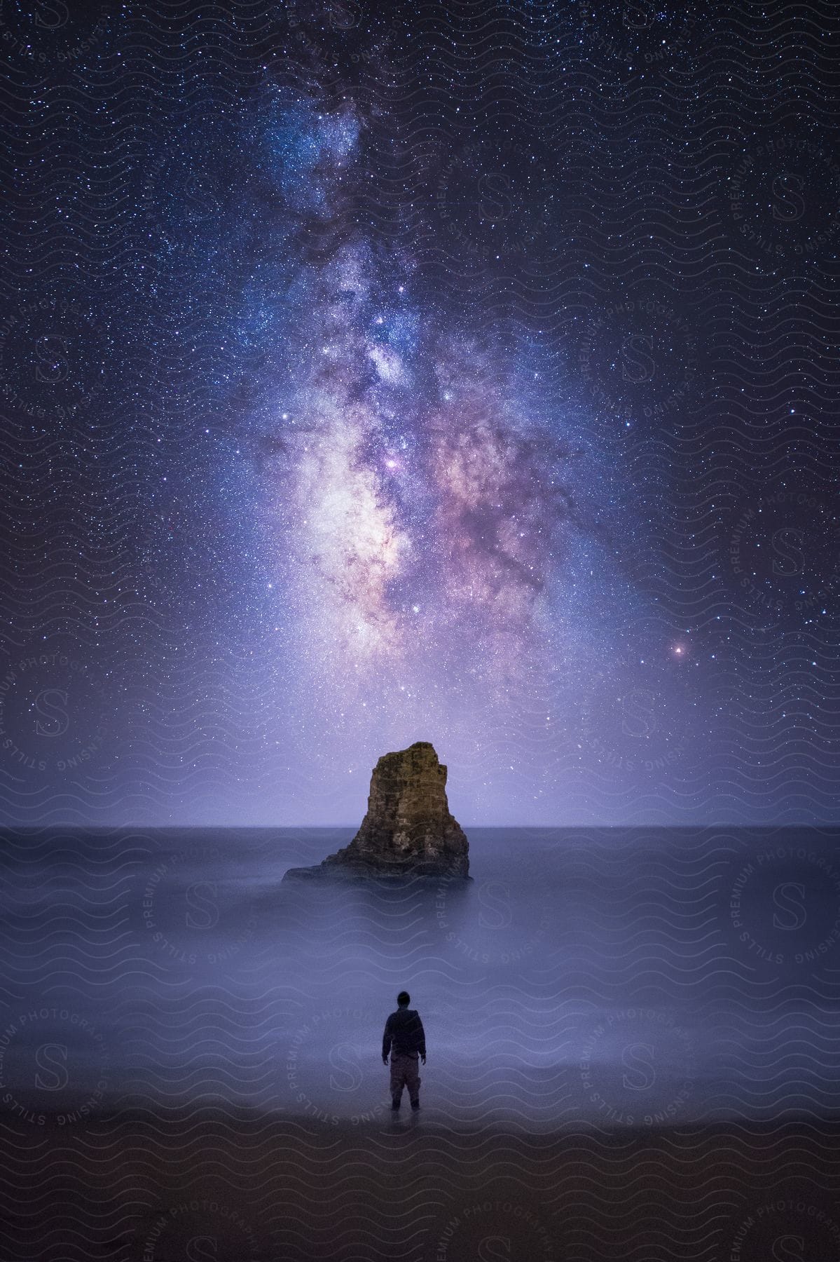 Man looks at the galaxy behind the rocky island in the foggy ocean.