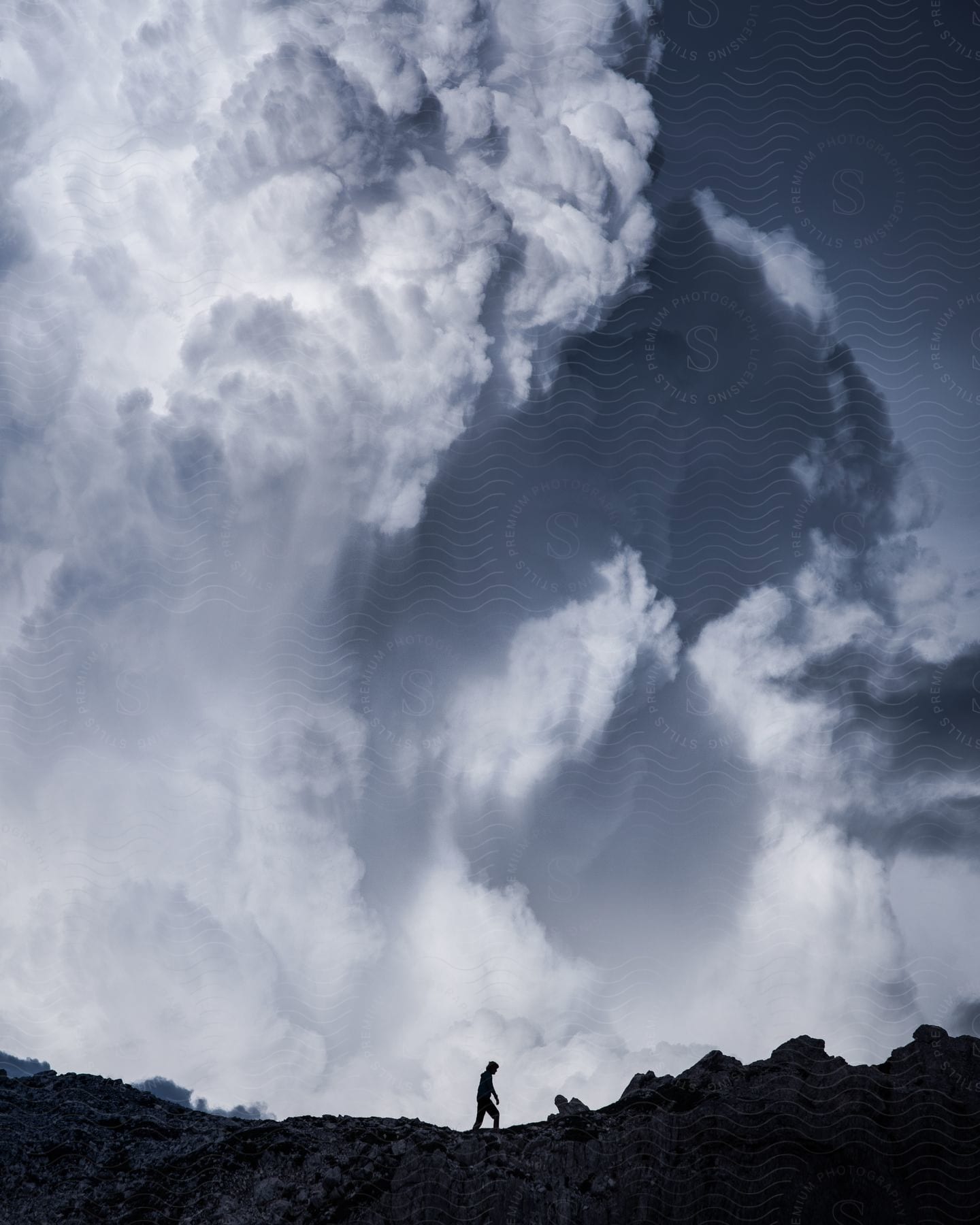 Silhouette of a person walking on a rocky mountain in a gray sky with neurotic white clouds.