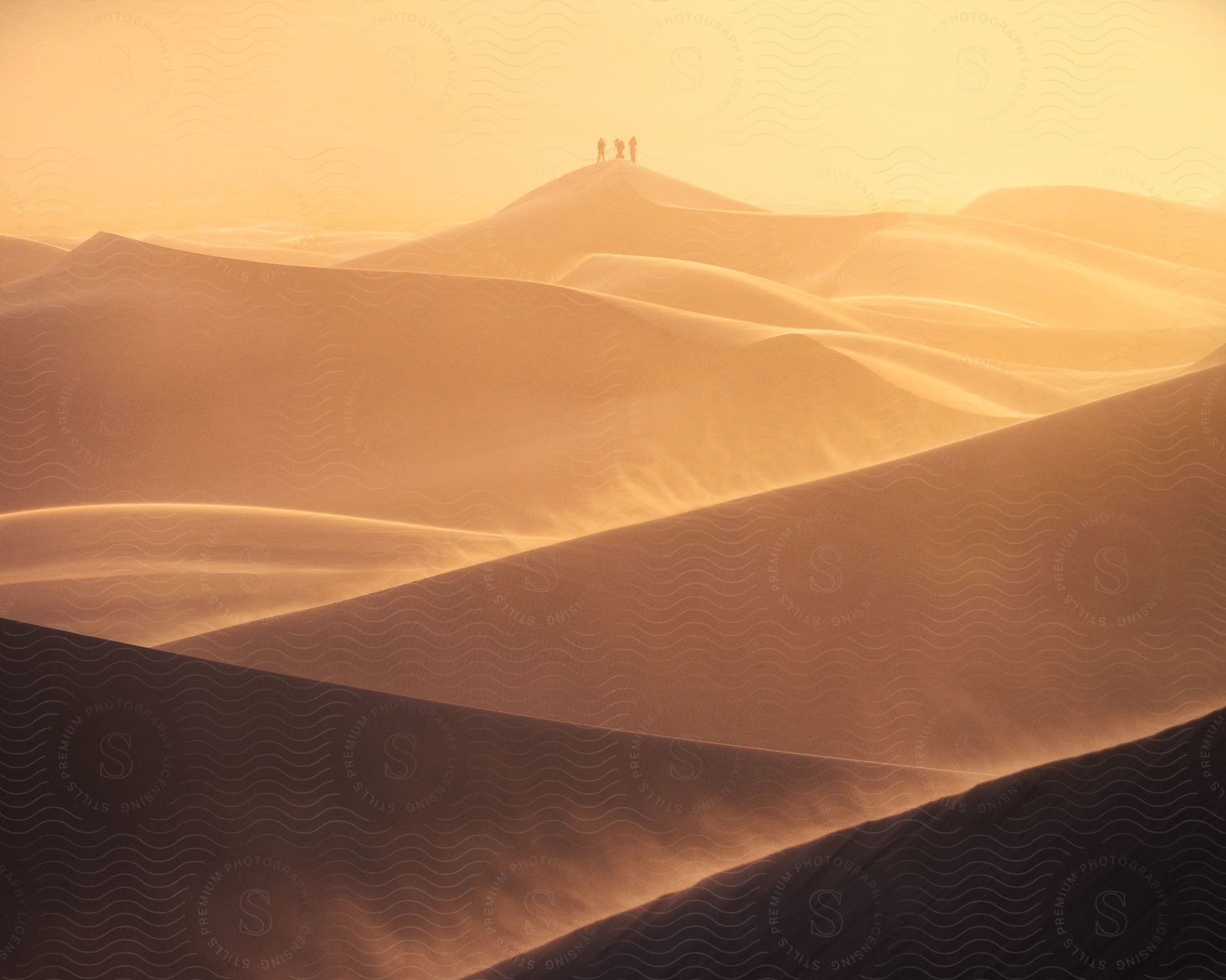 Stock photo of three figures were spotted on a tall sand dune in the distance on a windy day with blowing sand, with a yellow sky in the background.