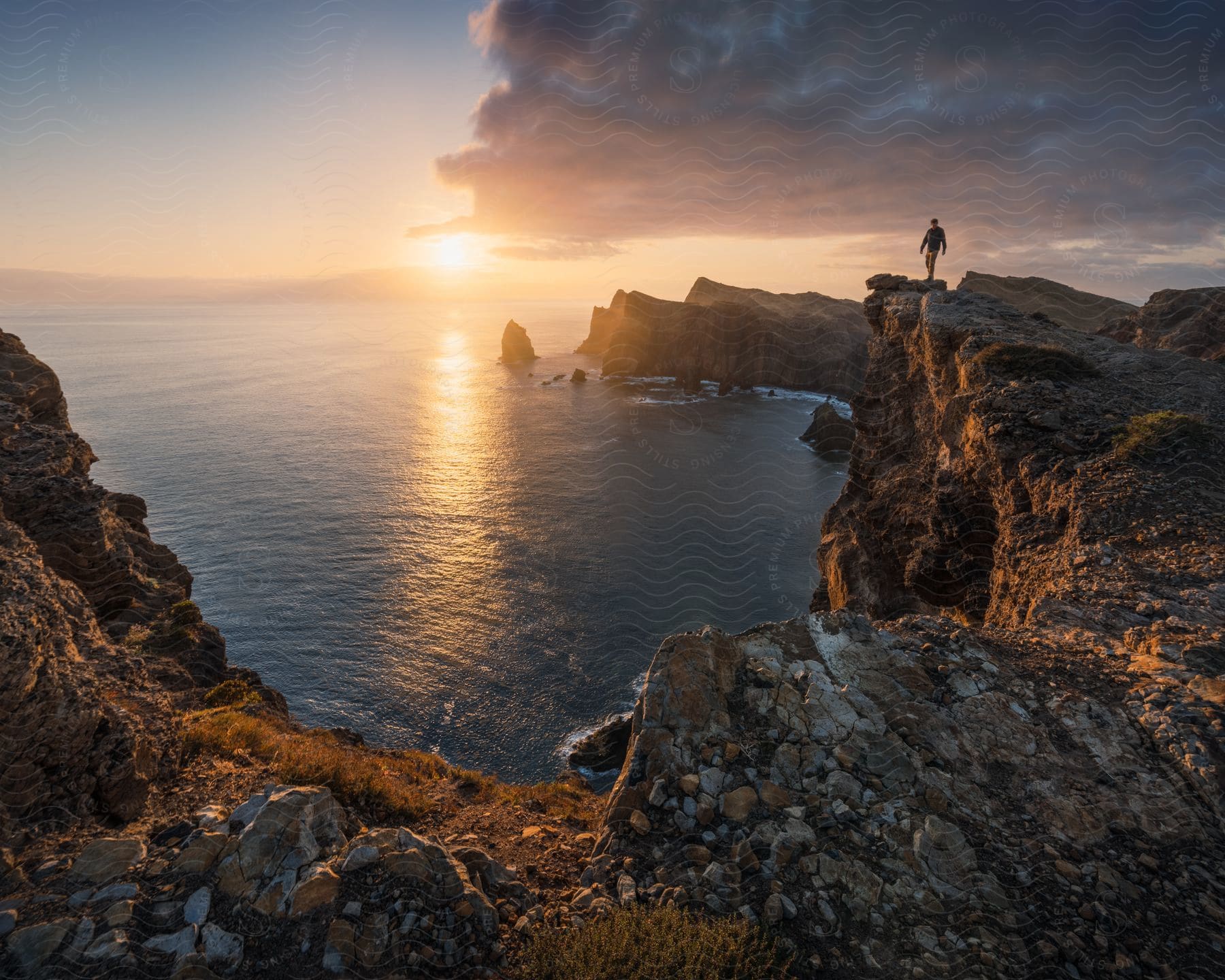 A person stands on a rocky coastline with the sun low in the sky