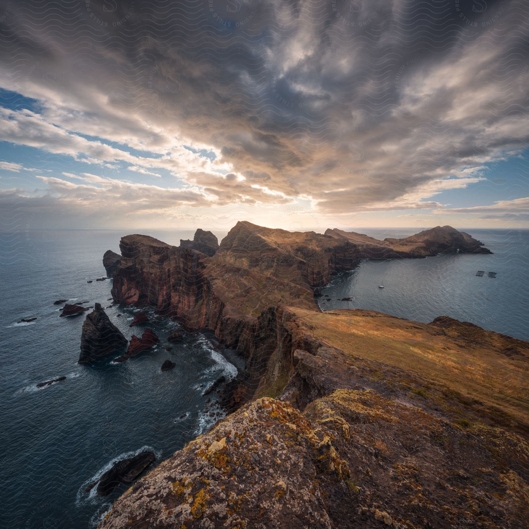 A rocky cliff overlooks a rocky coastline with water stretching to jagged mountain islands in the distance, all under cloudy skies.