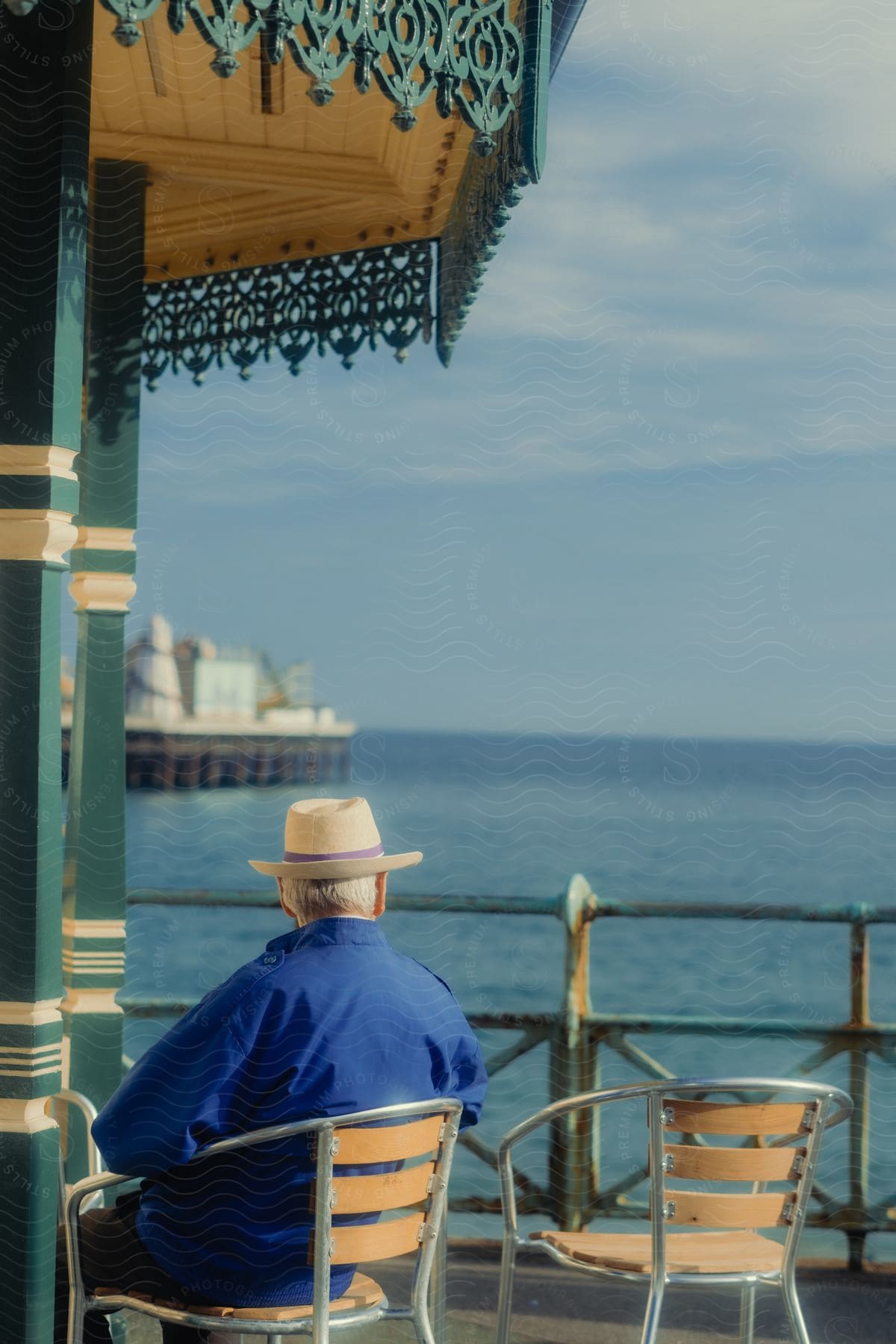 Man in a hat and blue shirt sitting on a bench on a pier, looking at the sea.