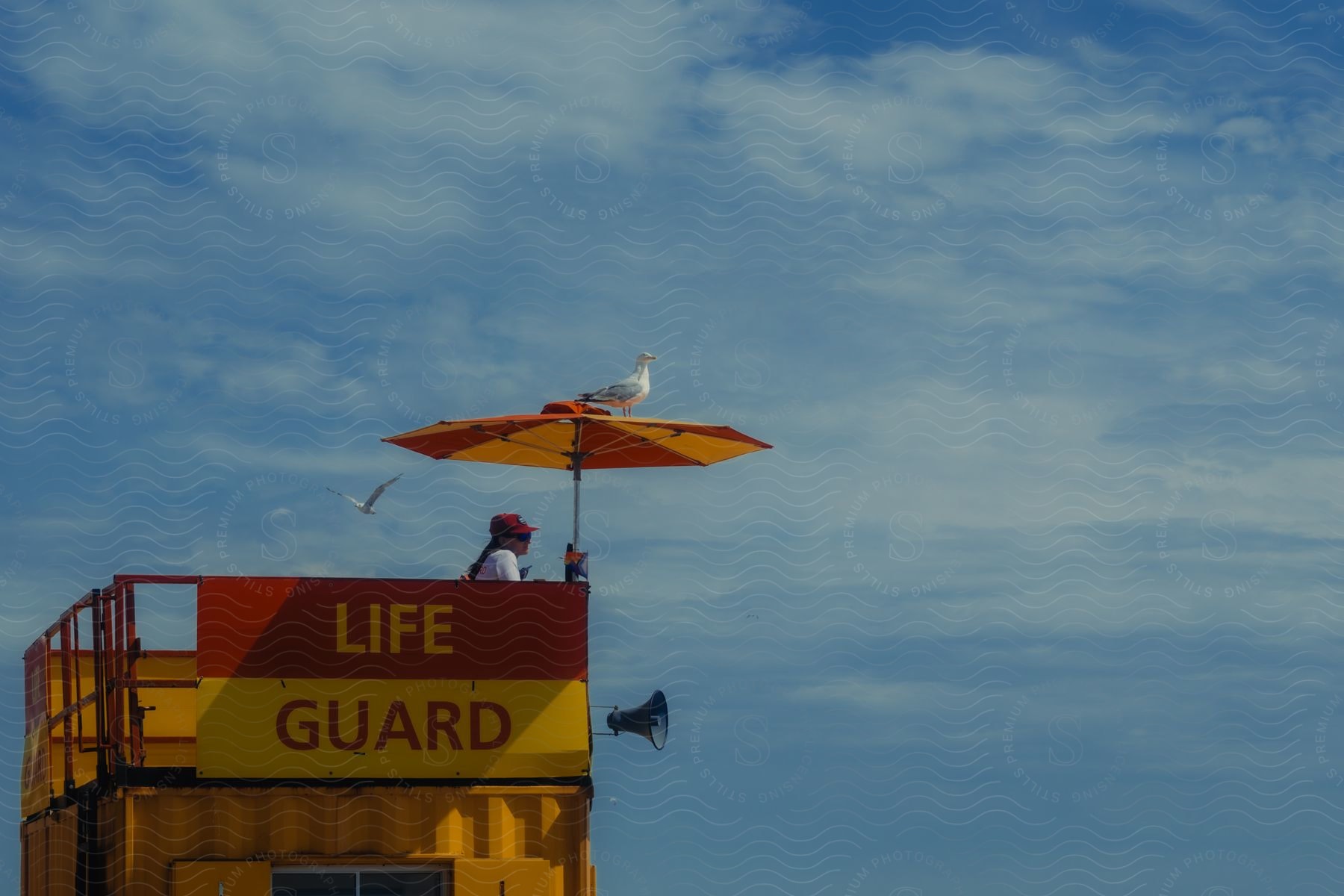 A lifeguard stands under a beach umbrella that's on top of a lifeguard house.