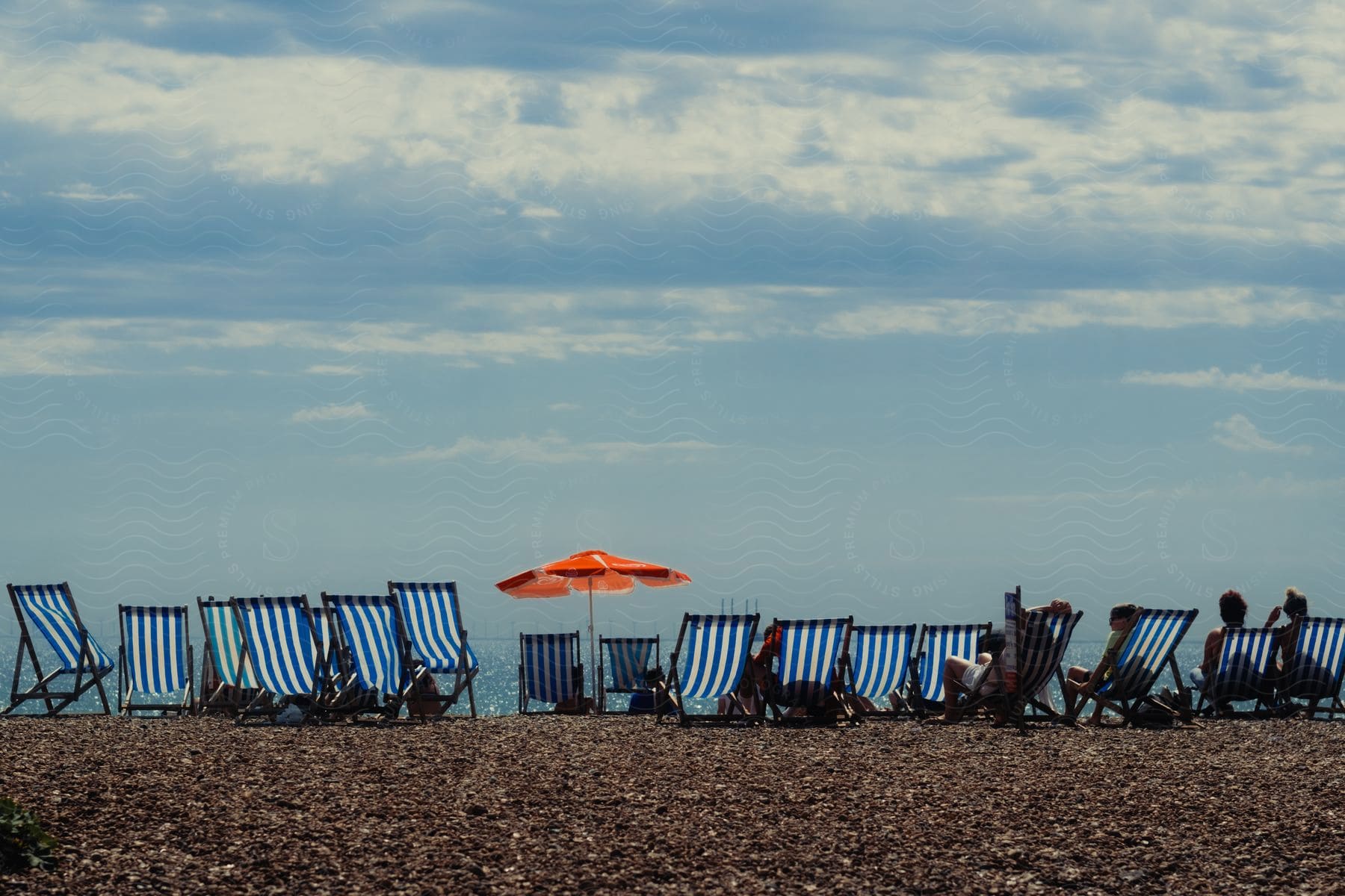 Row of blue striped deck chairs and a single orange umbrella on a beach with people sitting, looking out to sea under a cloudy sky.