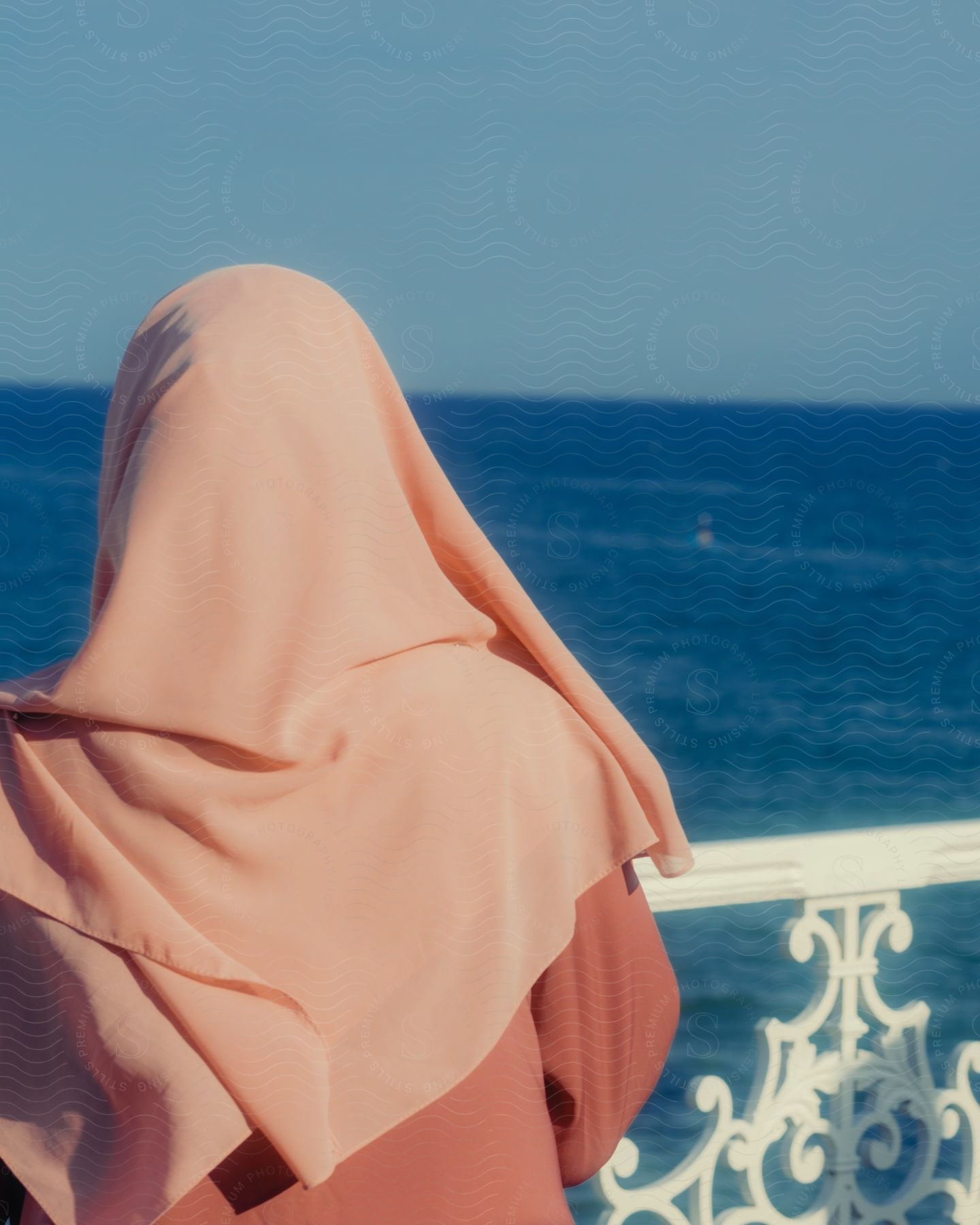 A person covered in a pink cloak looks at the blue sea. The scene conveys serenity and solitude, with the clear sky and vast ocean in front of you.