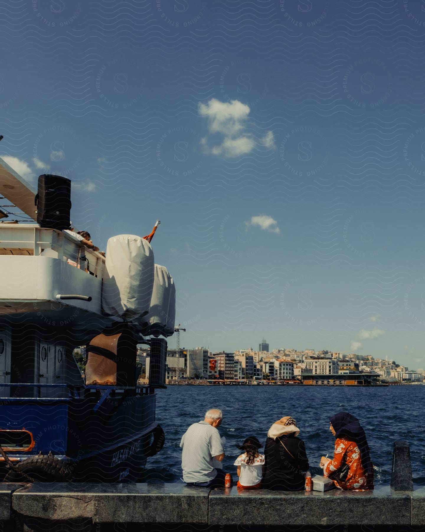 Seated people enjoy a moment by the sea, with a cityscape in the background and a boat to the side.