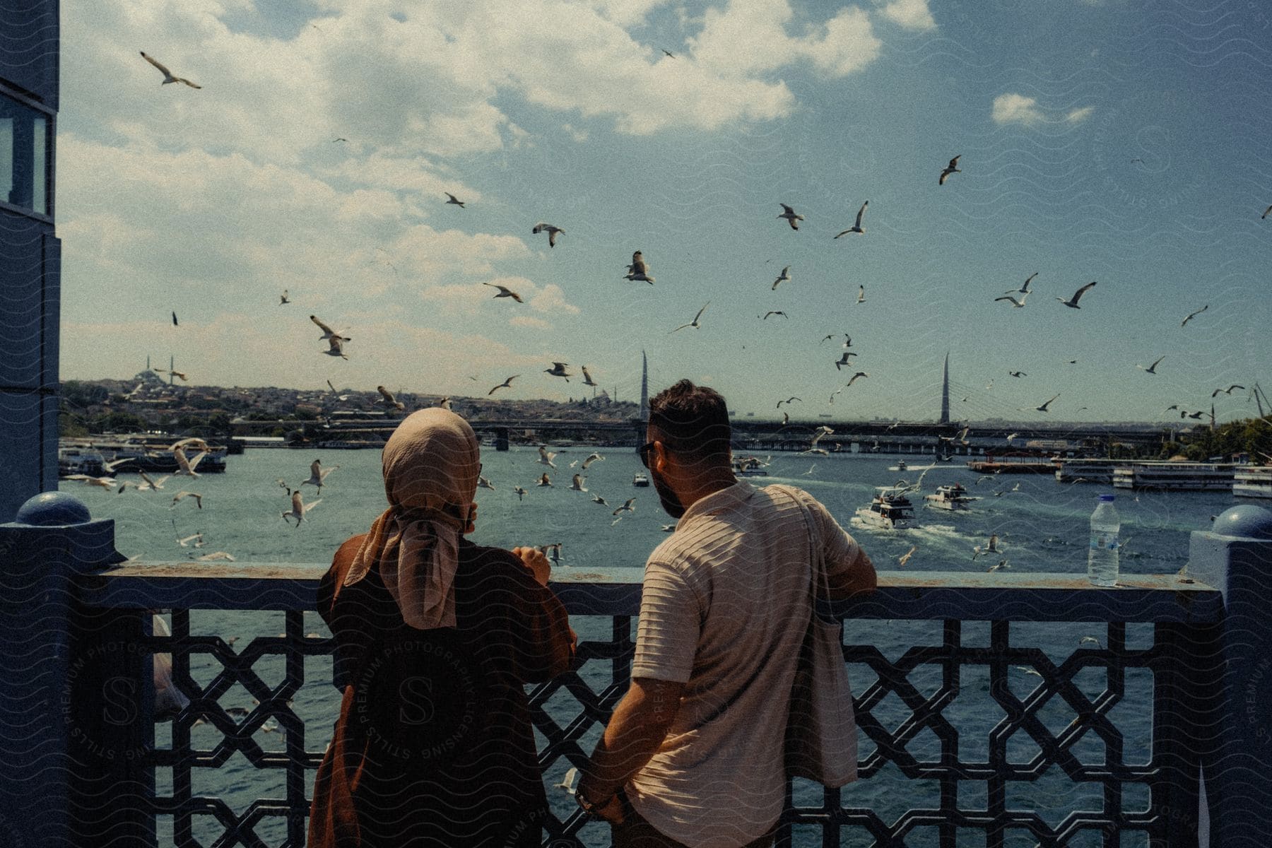 Man and woman back to back enjoying the view from a bridge to the sea with yachts and birds flying by during the day.