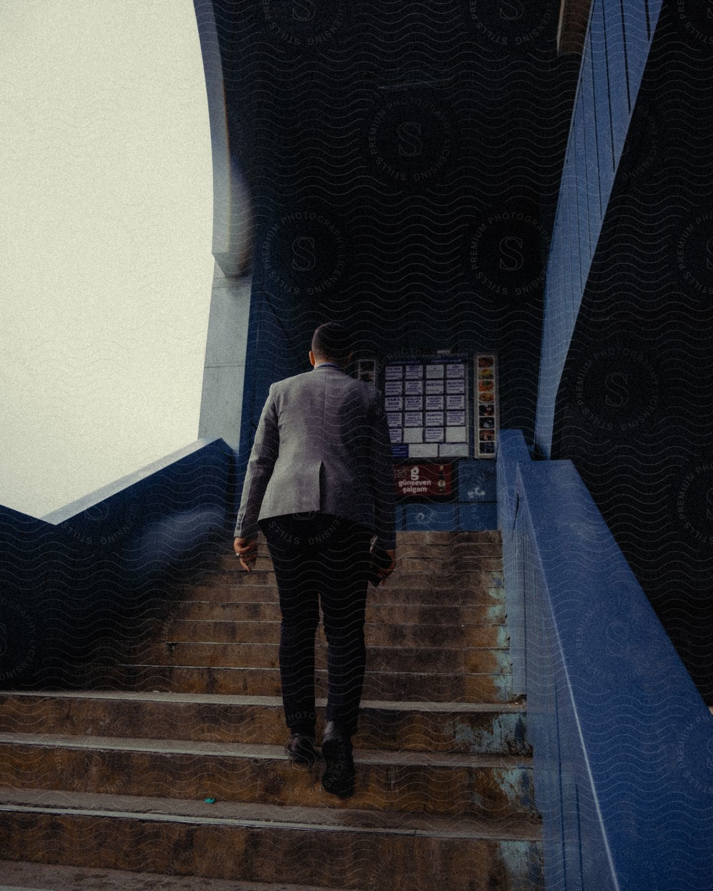 The person climbs a concrete staircase in a blue building, passing a wall with several posters.