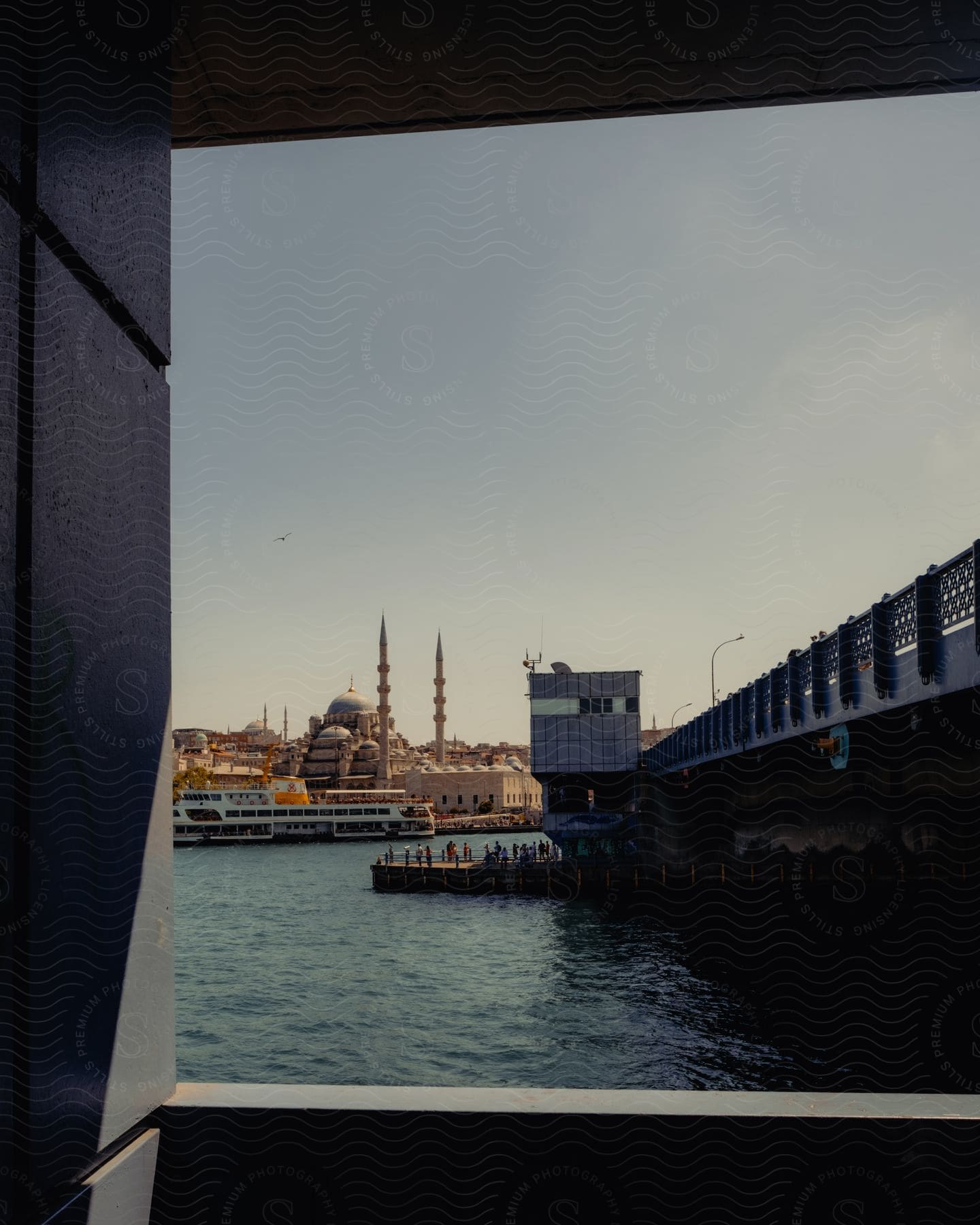 A boat is approaching and passing under a bridge, with a larger boat docked near the shore next to the bridge, all during the daytime.