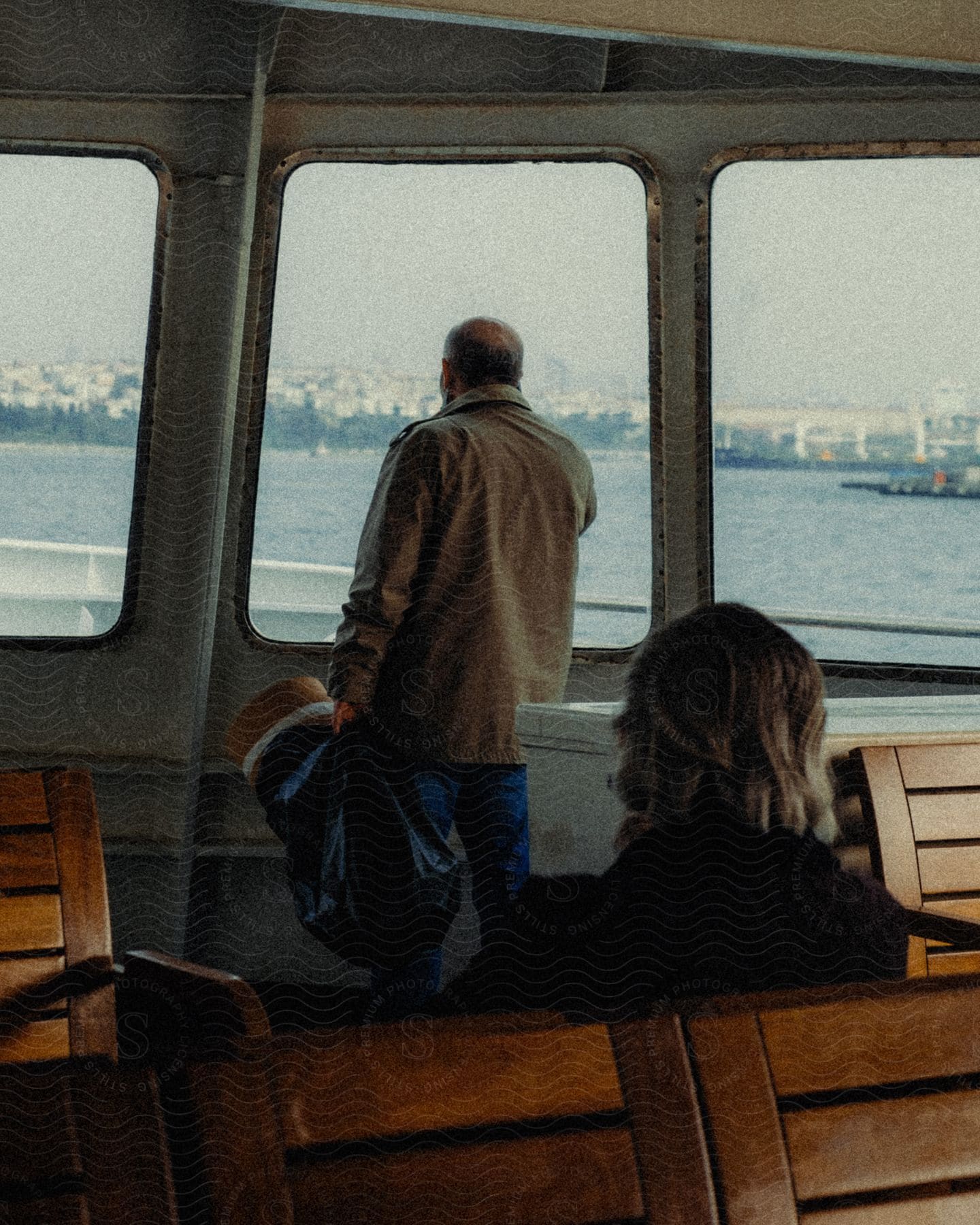 A person is standing on a boat, looking out of the window at the calm water and a city in the distance. The atmosphere is peaceful and contemplative.