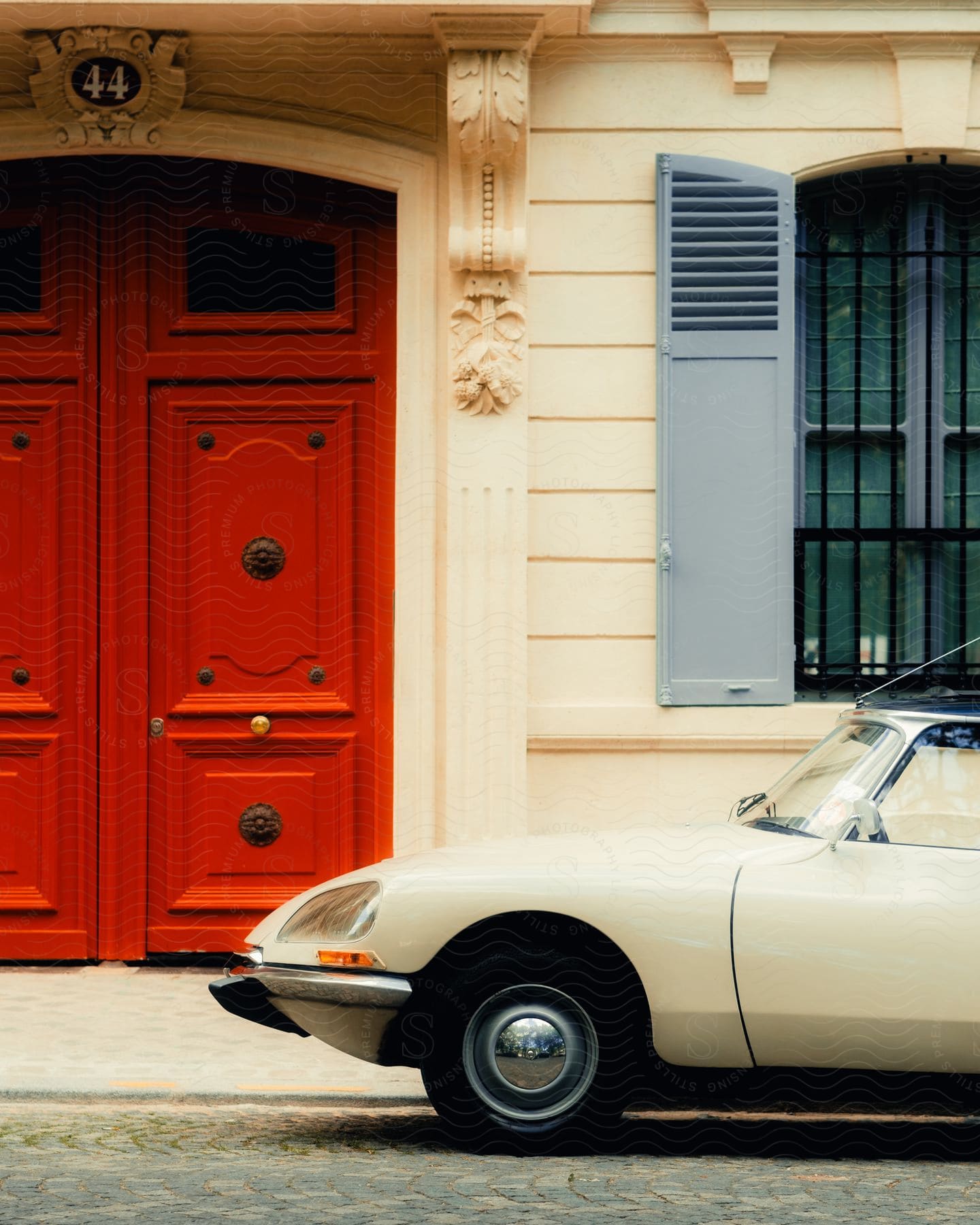 A small white car parked on the street in front of a building with a blue shutter window and red doors